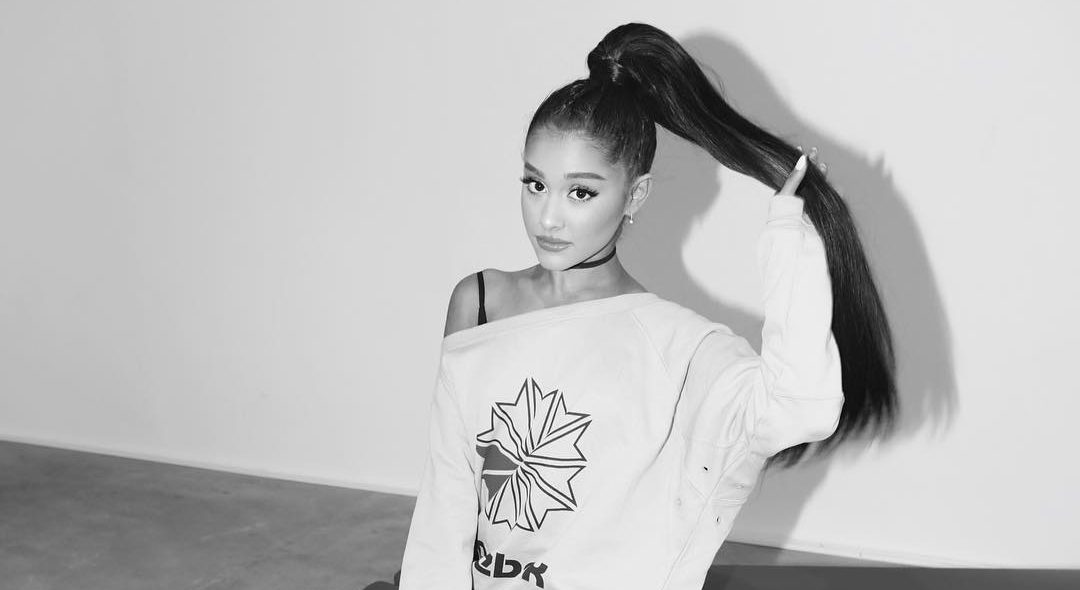 Ariana Grande com rabo de cavalo alto e moletom, sentada num sofá