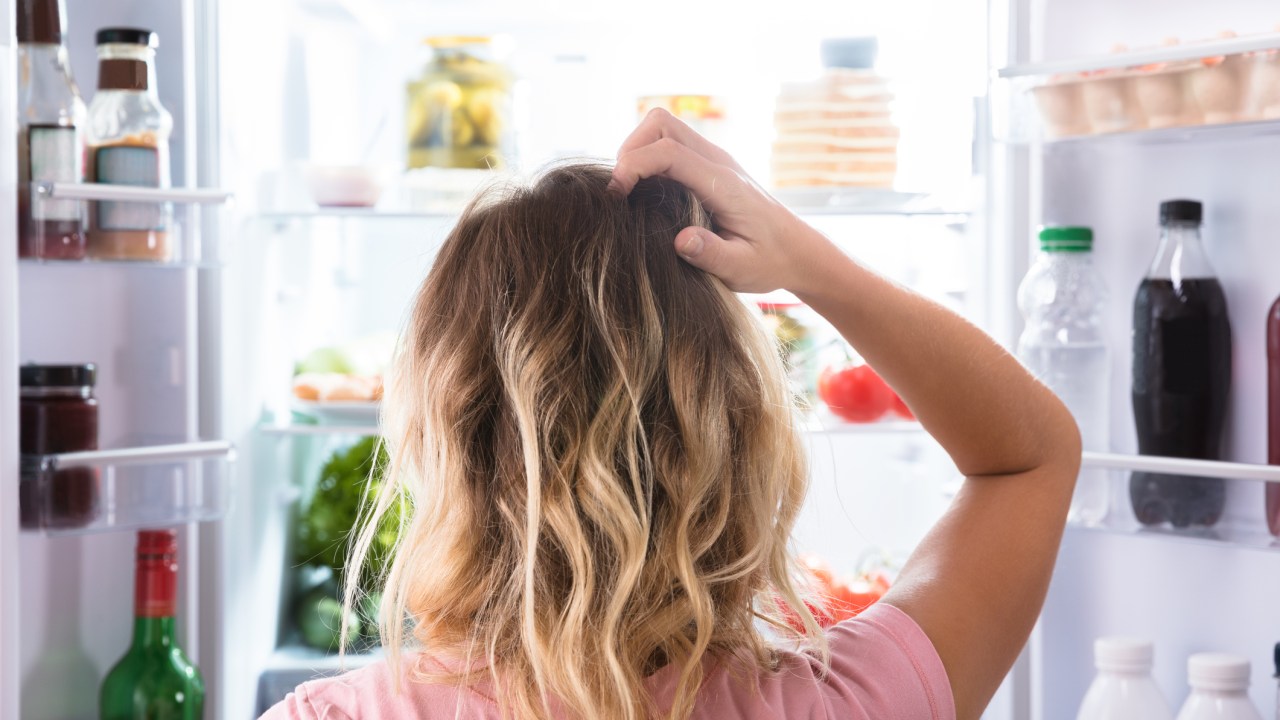 Mulher olhando para a geladeira com expressão de dúvida