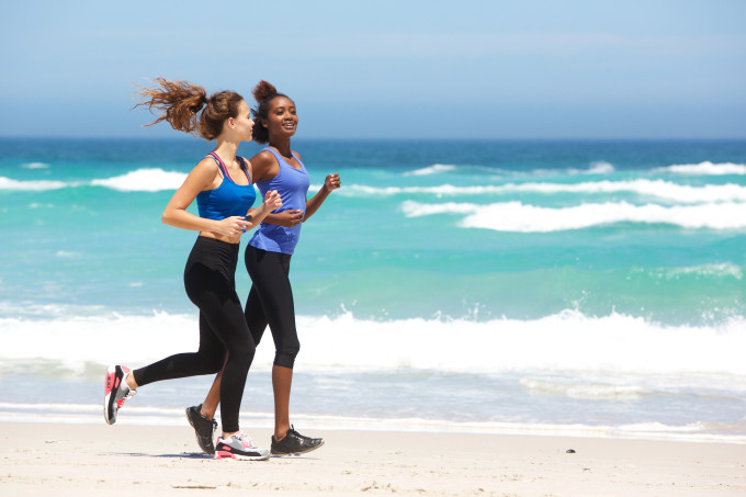 Duas mulheres correm na areia da praia e o mar está ao fundo.