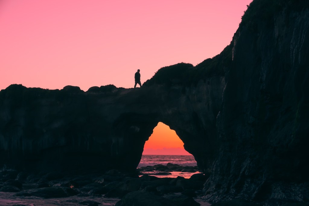 Foto de uma pedreira em cima ao mar, tem uma pessoa em cima e o céu ao fundo está rosa, como se fosse fim da tarde