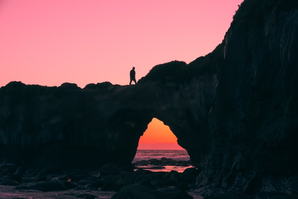 pexels-jeff-nissen-931911 Foto de uma pedreira em cima ao mar, tem uma pessoa em cima e o céu ao fundo está rosa, como se fosse fim da tarde