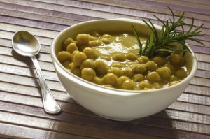 um bowl com sopa de lentilha e grão de bico e uma erva verde, sobre uma mesa de madeira e uma colher de metal posicionada ao lado