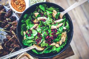 Salada de folhas verdes com frango em um recipiente escuro e talheres de metal posicionados dentro da tigela