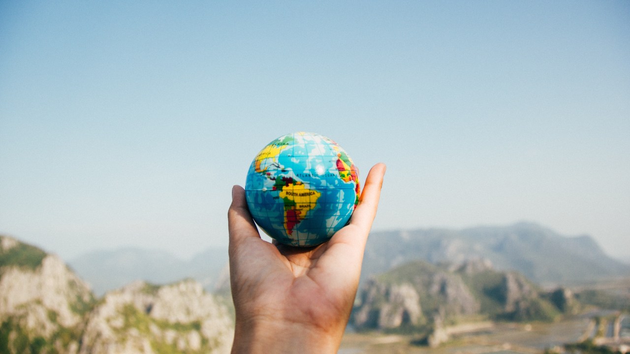 Mão segurando mini globo do mundo e fundo desfocado da natureza com montanhas