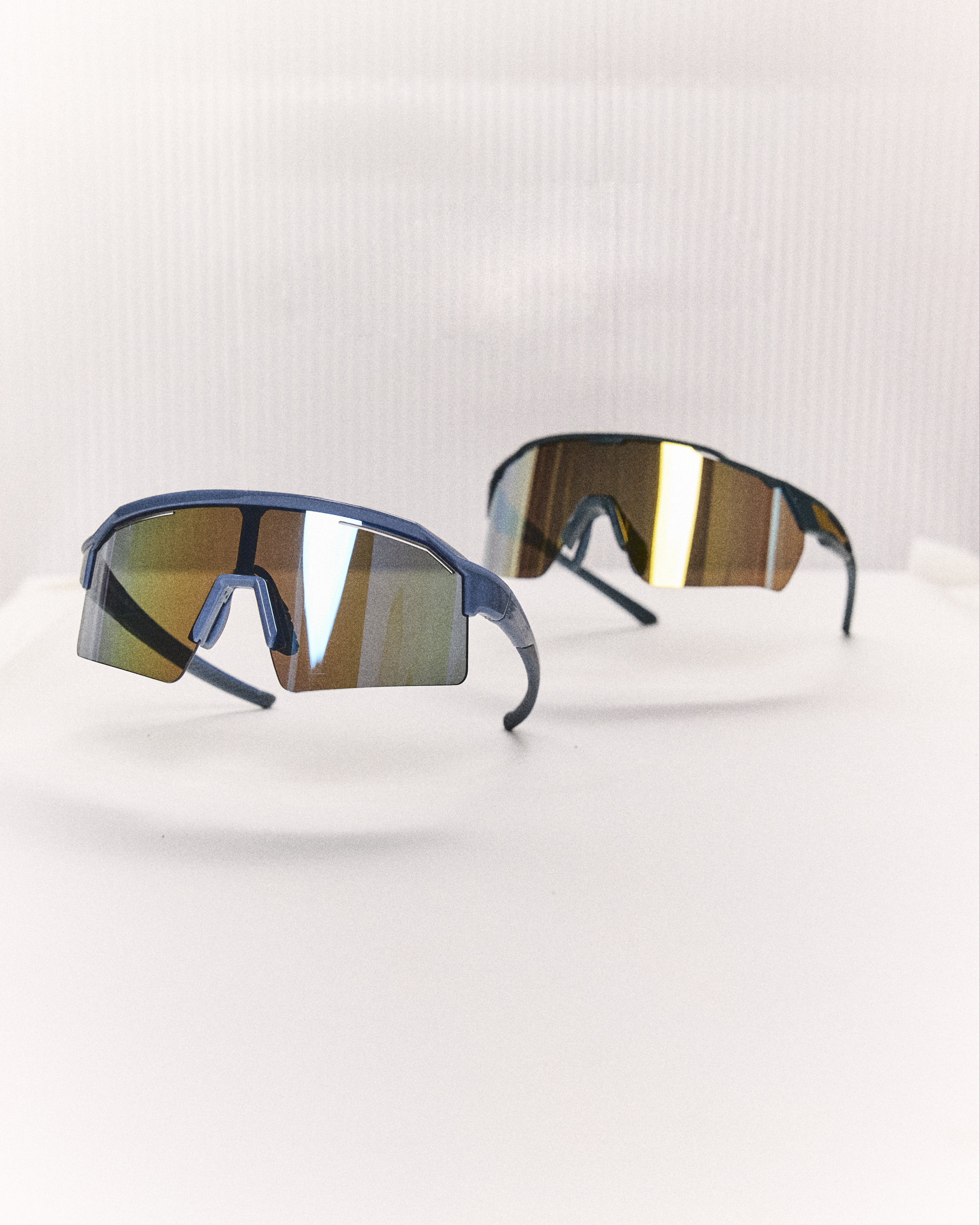 Dois &oacute;culos esportivos com lentes espelhadas coloridas, um azul escuro em primeiro plano e outro preto desfocado ao fundo, sobre uma superf&iacute;cie branca