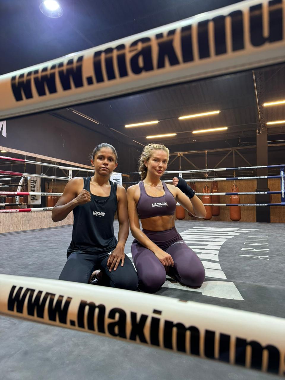 Duas mulheres, uma morena e uma loira, ajoelhadas em um ringue de boxe, vestindo roupas esportivas da marca Maximum. A morena usa regata e cal&ccedil;a preta, punho cerrado. A loira usa top e cal&ccedil;a roxa, com bandagens nas m&atilde;os. Ambas olham para frente, com o nome da marca vis&iacute;vel nas cordas do ringue e ao fundo. Sacos de pancada pendurados completam o cen&aacute;rio da academia