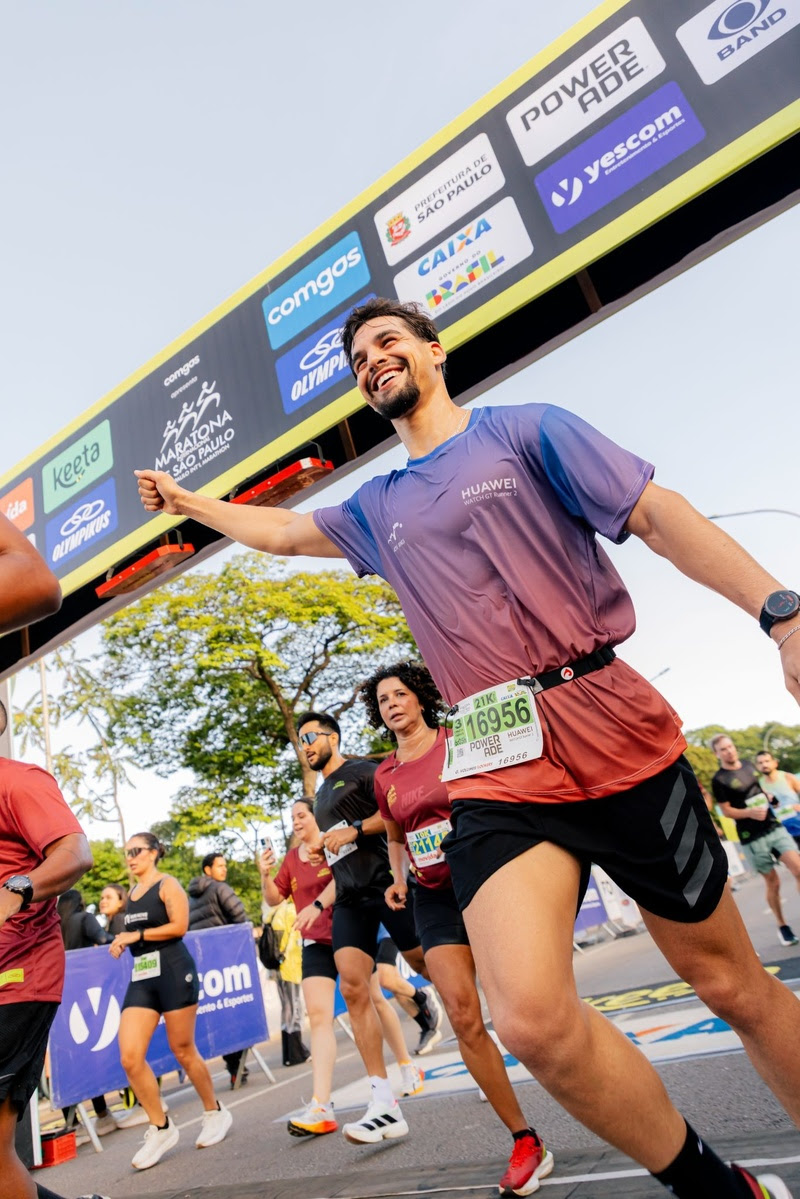 Corredor sorridente com camiseta roxa e vermelha, shorts pretos, bra&ccedil;o estendido e rel&oacute;gio esportivo, cruzando a linha de chegada de uma maratona. Outros corredores e um banner de patrocinadores ao fundo, sob c&eacute;u claro