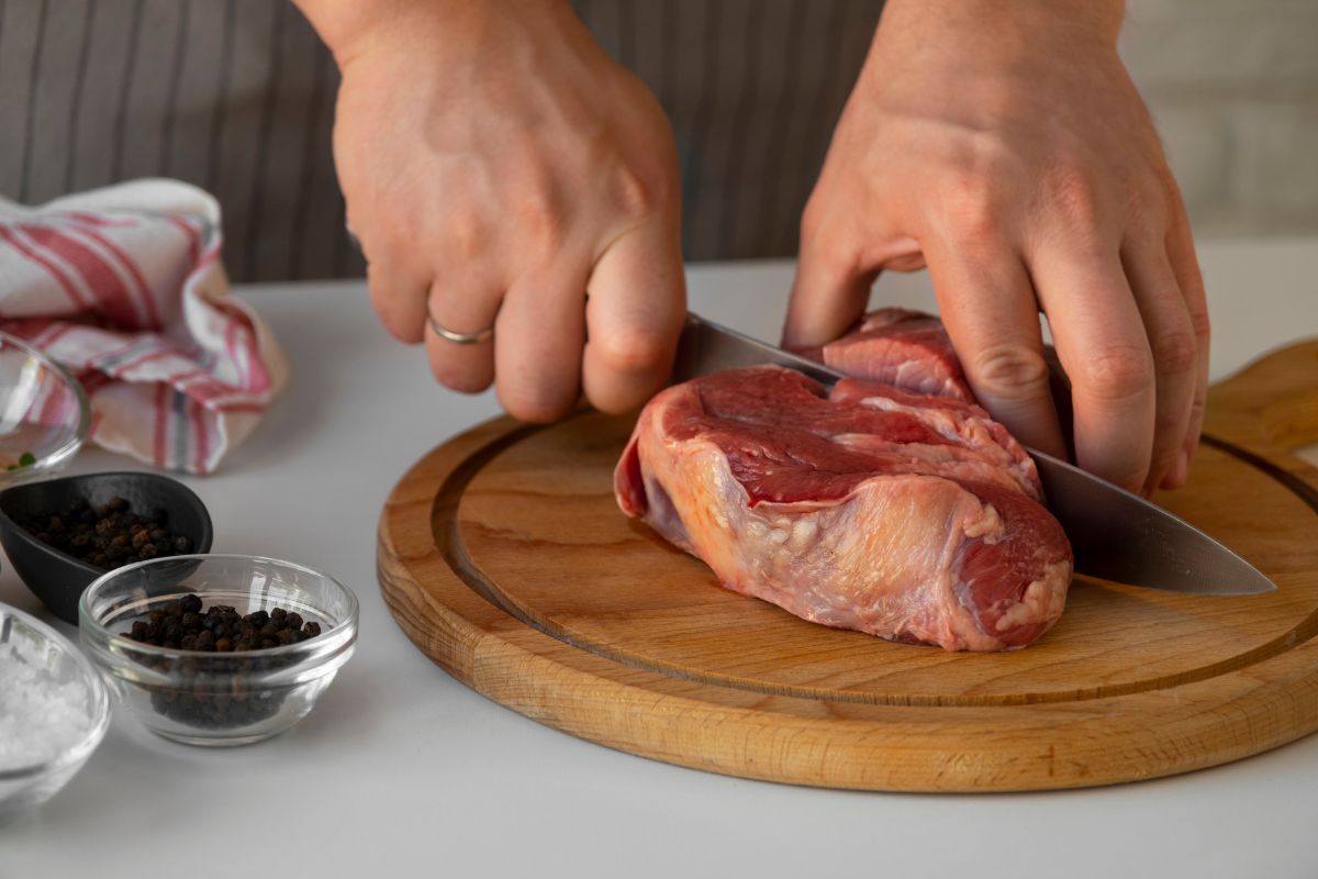 Mãos de uma pessoa cortando um pedaço de carne vermelha crua com uma faca em uma tábua de madeira, com potes de sal e pimenta ao lado