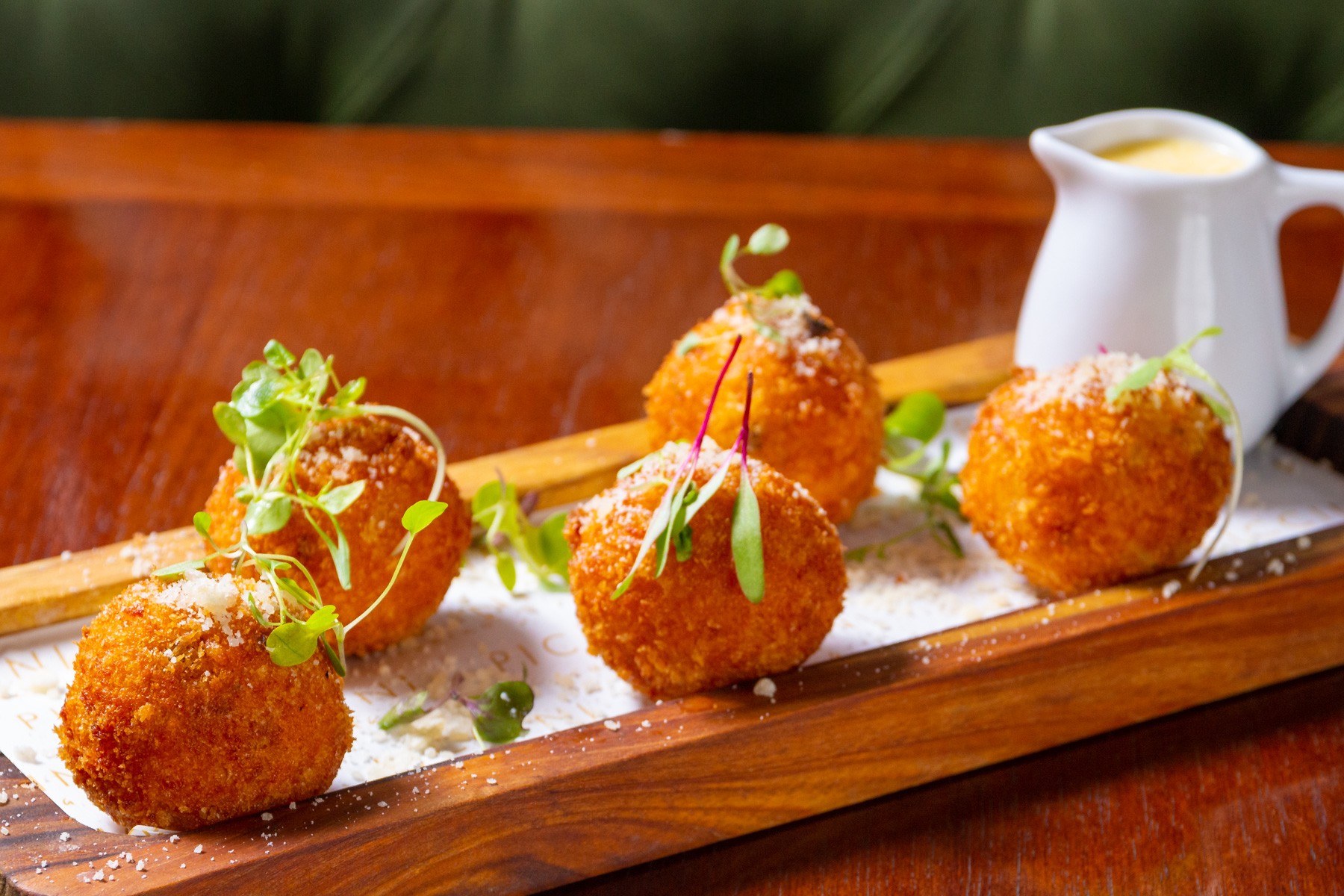 Cinco bolinhos fritos dourados, polvilhados com queijo ralado e decorados com brotos verdes, servidos em uma t&aacute;bua de madeira com um molho cremoso em jarra branca