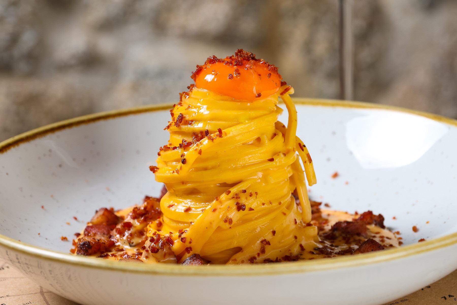 Prato de espaguete &agrave; carbonara, com massa amarela enrolada em torre, gema de ovo crua no topo e peda&ccedil;os de bacon crocante espalhados, servido em um prato branco com borda dourada