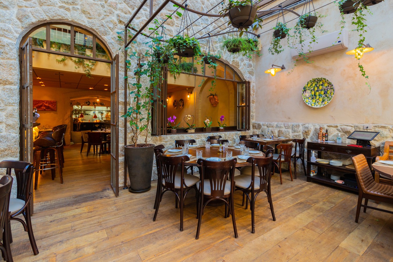 P&aacute;tio de restaurante com piso de madeira, paredes de pedra e plantas penduradas. H&aacute; mesas redondas e quadradas com cadeiras de madeira, prontas para refei&ccedil;&otilde;es. Uma porta arqueada aberta revela o interior do restaurante, com balc&atilde;o e cozinha ao fundo. Uma janela com orqu&iacute;deas e um prato decorativo de lim&otilde;es adornam a parede.