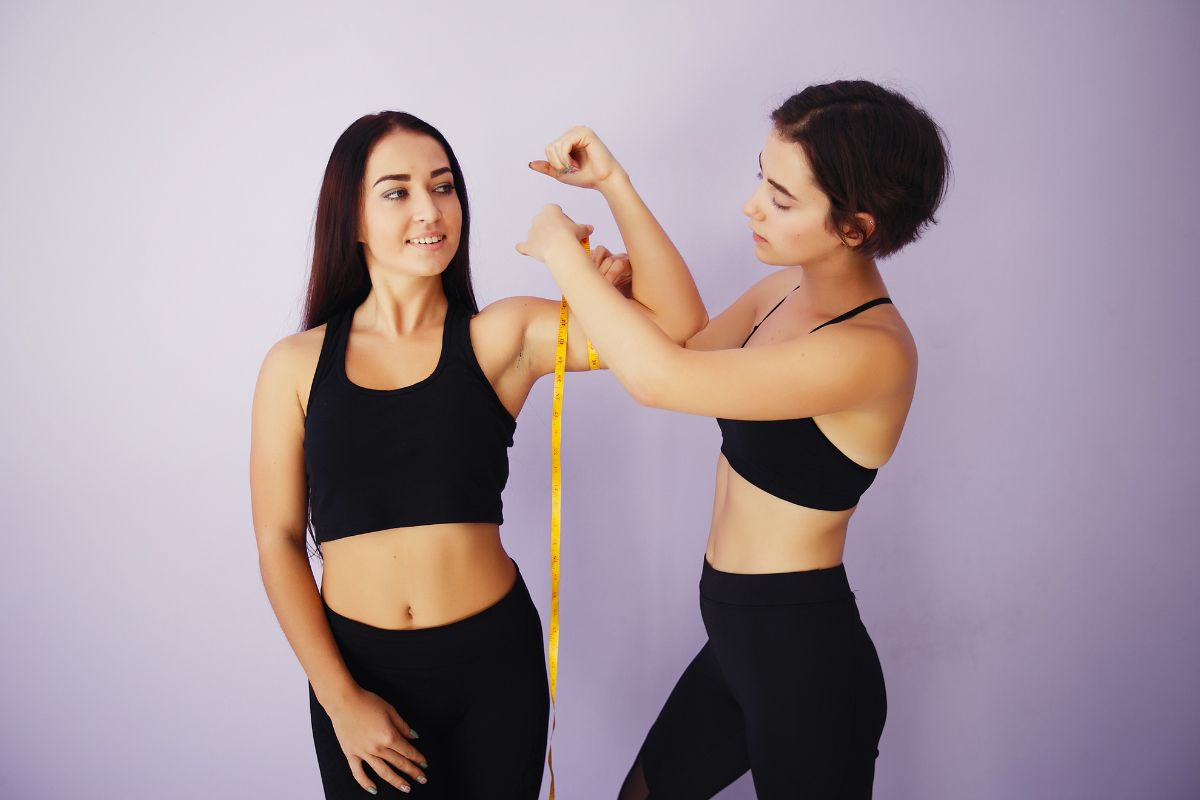 Duas mulheres jovens, vestindo roupas de ginástica pretas, em um fundo lilás. Uma mede o bíceps flexionado da outra com uma fita métrica amarela, enquanto a outra sorri olhando para o lado