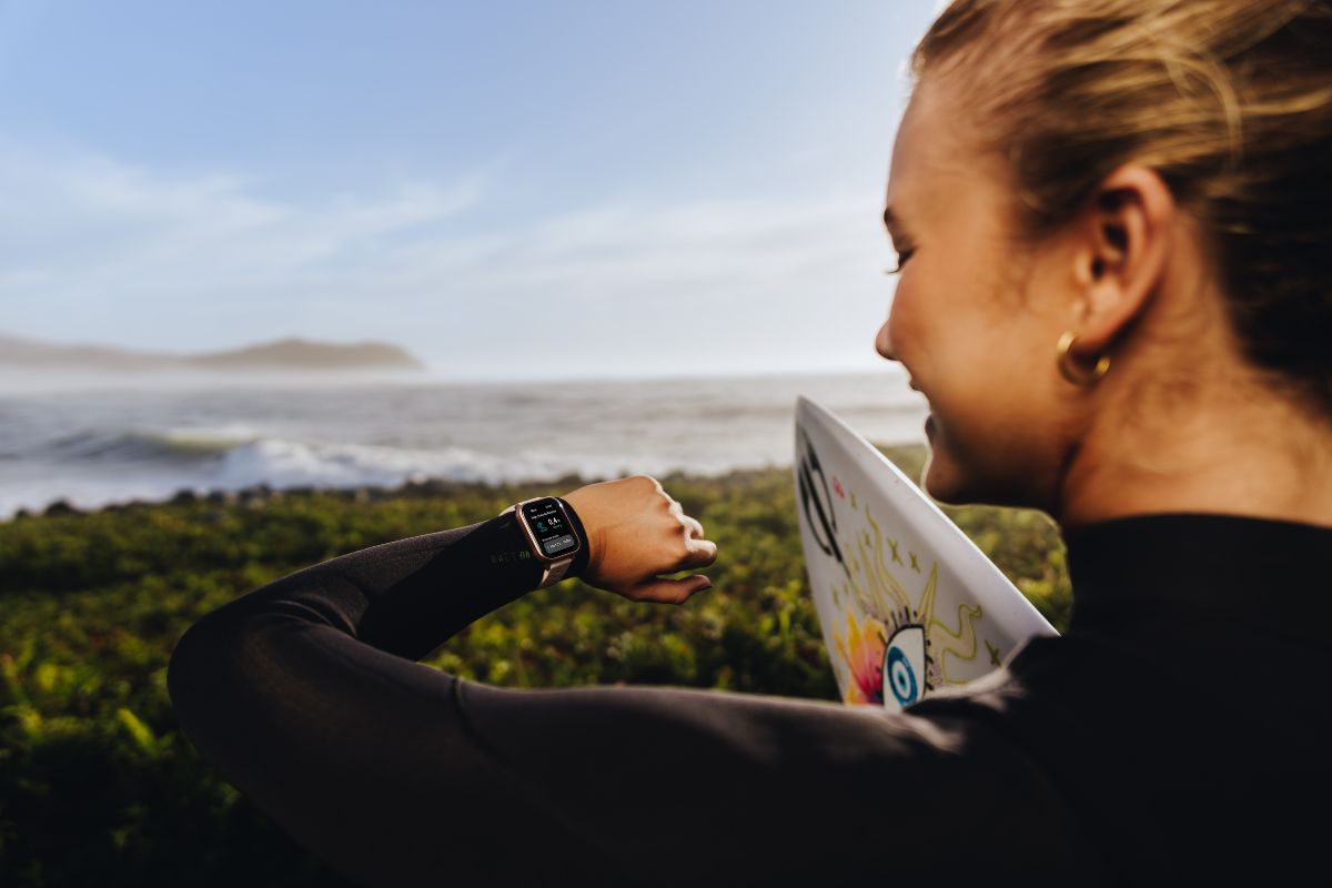 Mulher sorridente de perfil, vestindo roupa de neoprene preta, olhando um smartwatch no pulso esquerdo. Ela segura uma prancha de surfe branca com desenhos coloridos, em frente a um mar com ondas e vegeta&ccedil;&atilde;o verde sob c&eacute;u azul