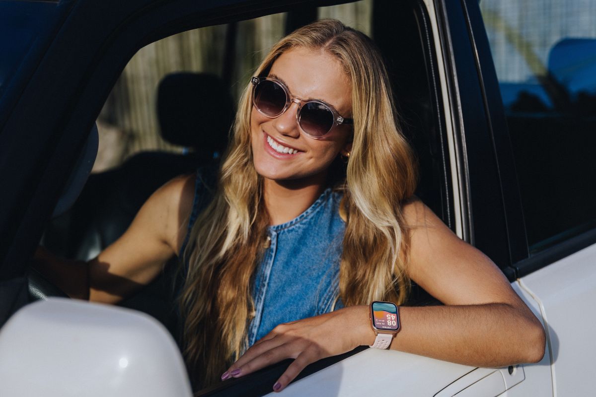 Mulher loira sorridente, com &oacute;culos de sol redondos e blusa jeans, apoiada na janela de um carro branco, exibindo um smartwatch rosa no pulso