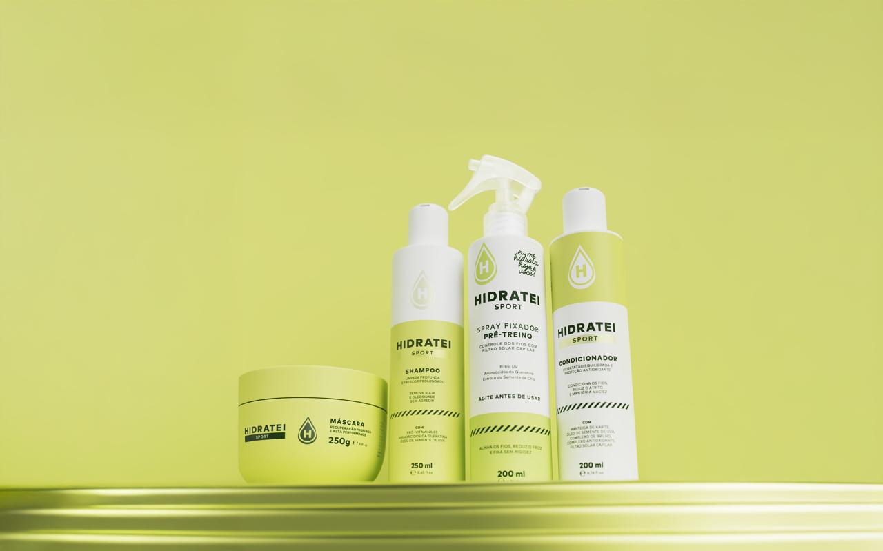 Quatro produtos da linha Hidratei Sport em embalagens brancas e verde-limão sobre uma prateleira dourada, com fundo verde-limão. Da esquerda para a direita: um pote de máscara capilar de 250g, um frasco de shampoo de 250ml, um spray fixador pré-treino de 200ml e um condicionador de 200ml
