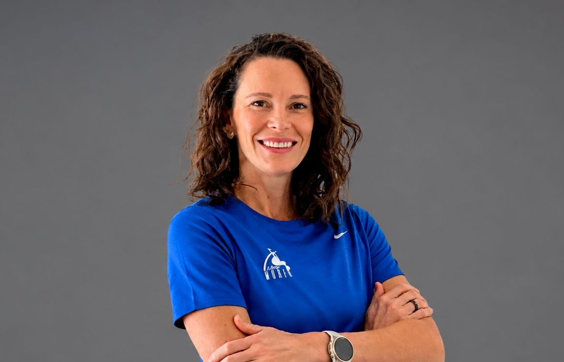 Mulher de cabelos castanhos cacheados sorri, vestindo camiseta azul com logo branco e relógio esportivo prateado no pulso esquerdo, braços cruzados sobre fundo cinza