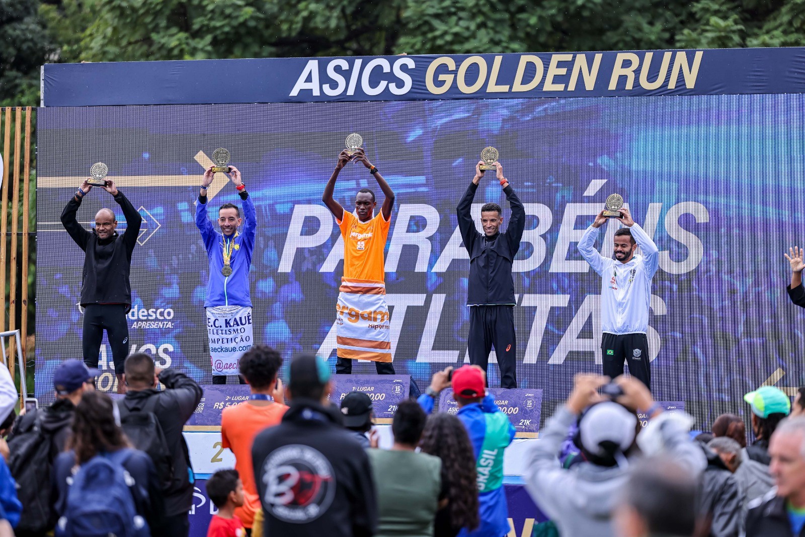 Cinco atletas masculinos sorridentes no p&oacute;dio da Asics Golden Run, erguendo trof&eacute;us. O vencedor, ao centro, veste laranja, os demais usam roupas esportivas escuras e claras. Uma multid&atilde;o os observa e fotografa