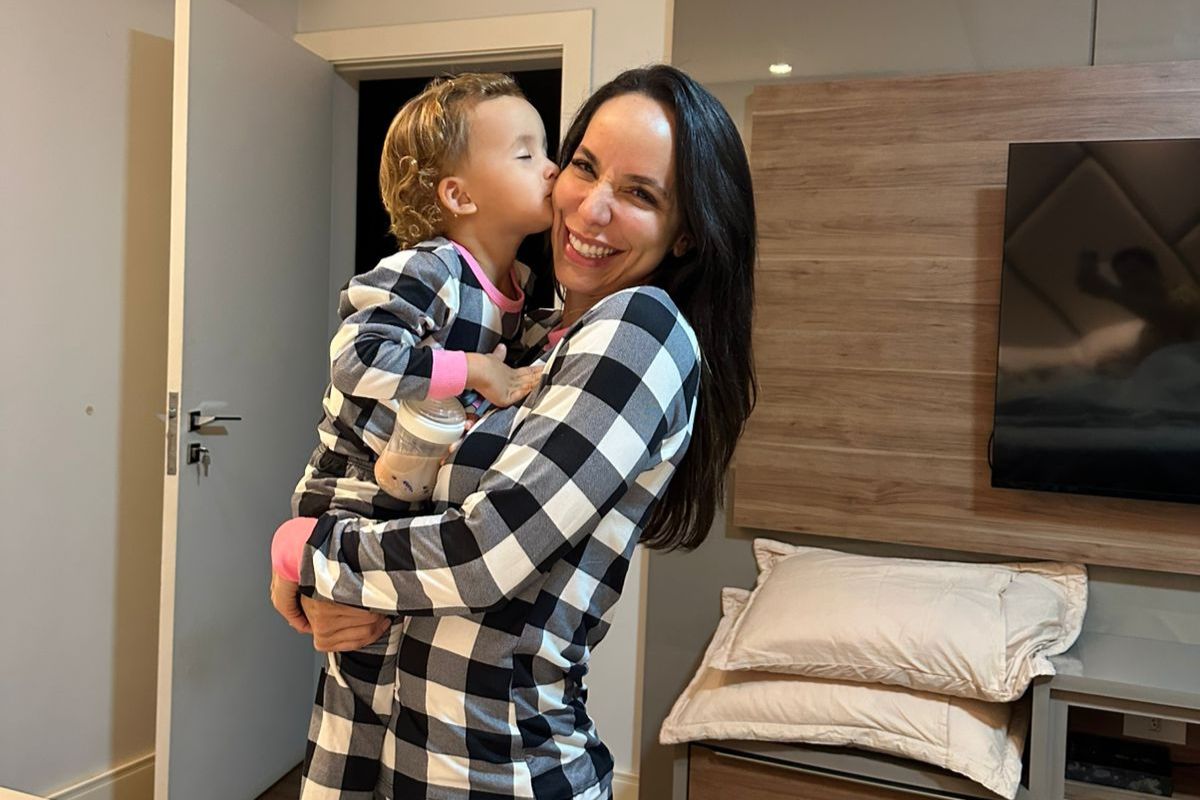 Mulher de cabelos escuros e crian&ccedil;a loira, ambas de pijama xadrez preto e branco, se abra&ccedil;am. A crian&ccedil;a beija a bochecha da mulher, que sorri. A crian&ccedil;a segura uma mamadeira. Ao fundo, uma porta e um painel de madeira com televis&atilde;o
