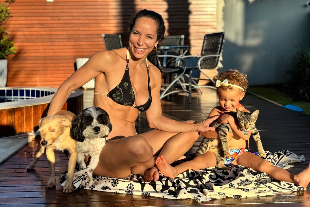 Mulher sorridente de biqu&iacute;ni preto e uma crian&ccedil;a com la&ccedil;o amarelo na cabe&ccedil;a sentadas em um deck de madeira, com dois cachorros e um gato listrado, sob luz solar