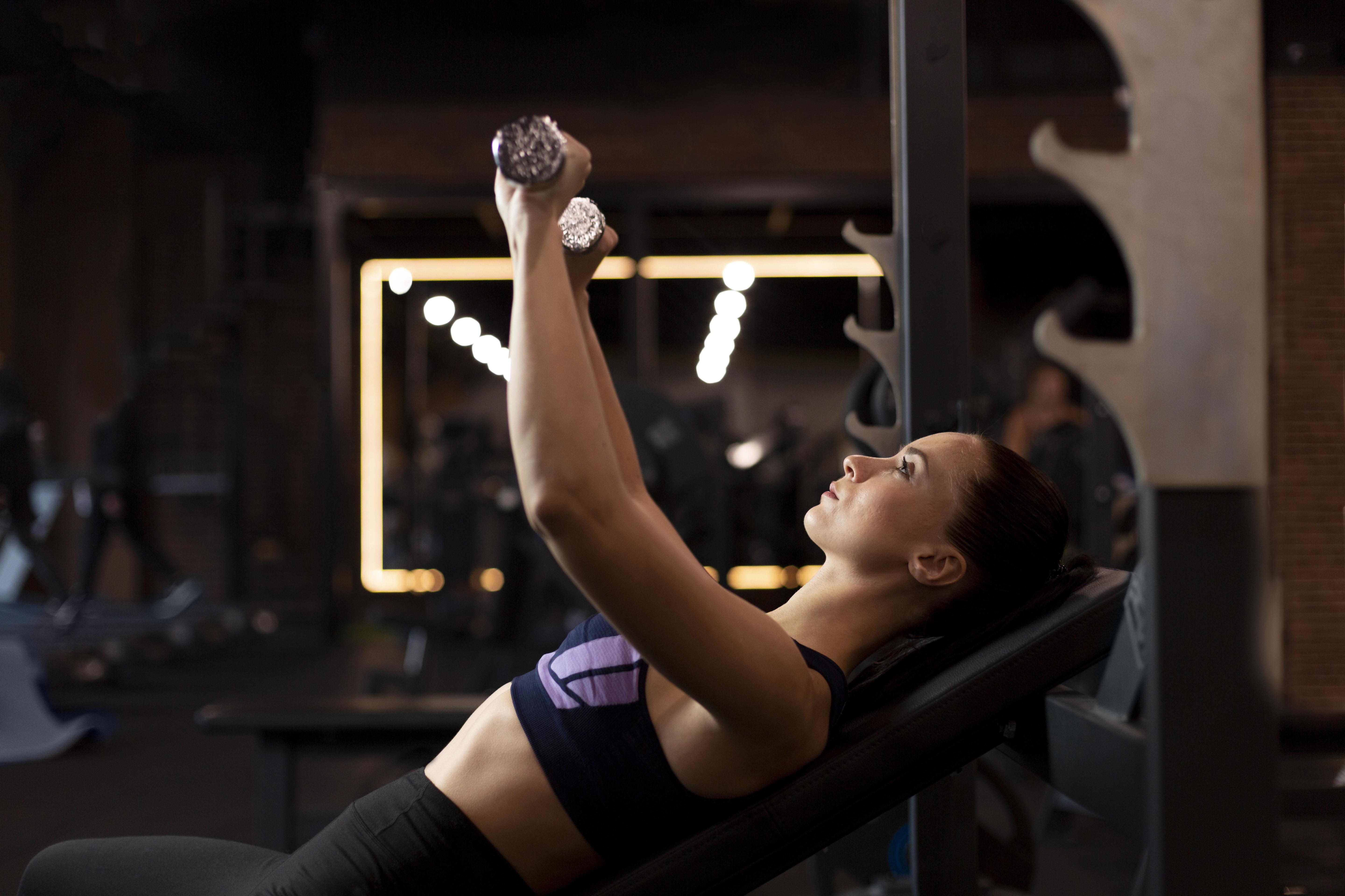 Mulher de perfil, deitada em banco inclinado na academia, levantando halteres com os braços estendidos para cima, vestindo top esportivo roxo e preto