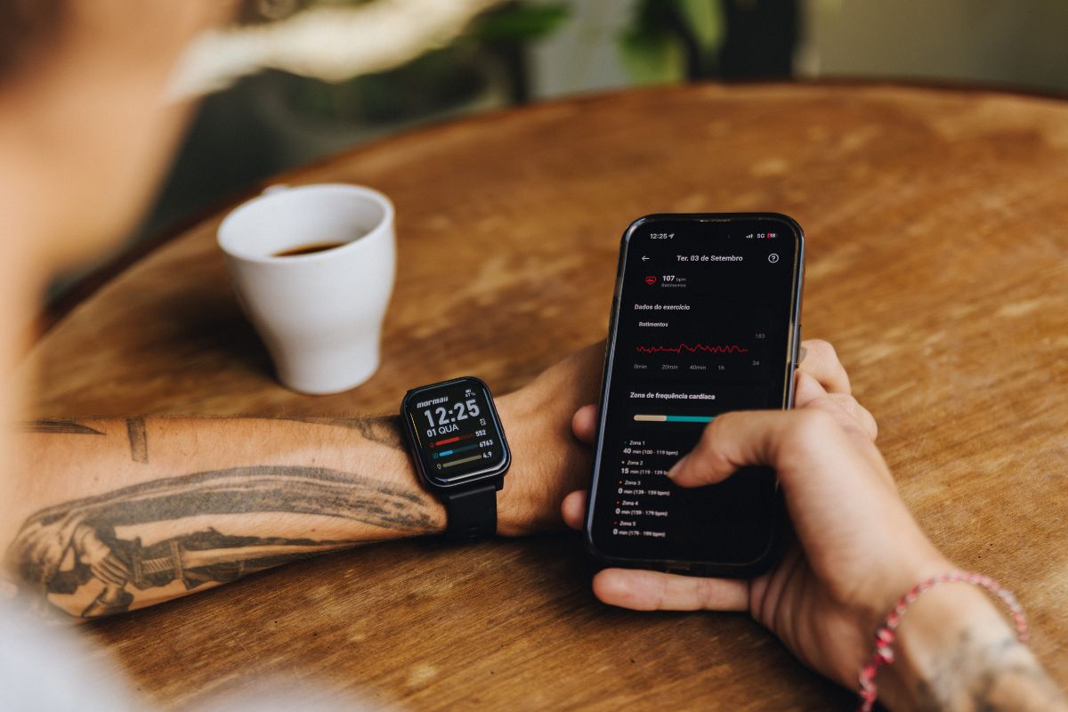 Pessoa com bra&ccedil;o tatuado e rel&oacute;gio inteligente preto no pulso esquerdo, segurando um smartphone com a m&atilde;o direita. O celular exibe dados de exerc&iacute;cio e frequ&ecirc;ncia card&iacute;aca em tela escura. Uma x&iacute;cara branca de caf&eacute; est&aacute; sobre a mesa de madeira ao lado