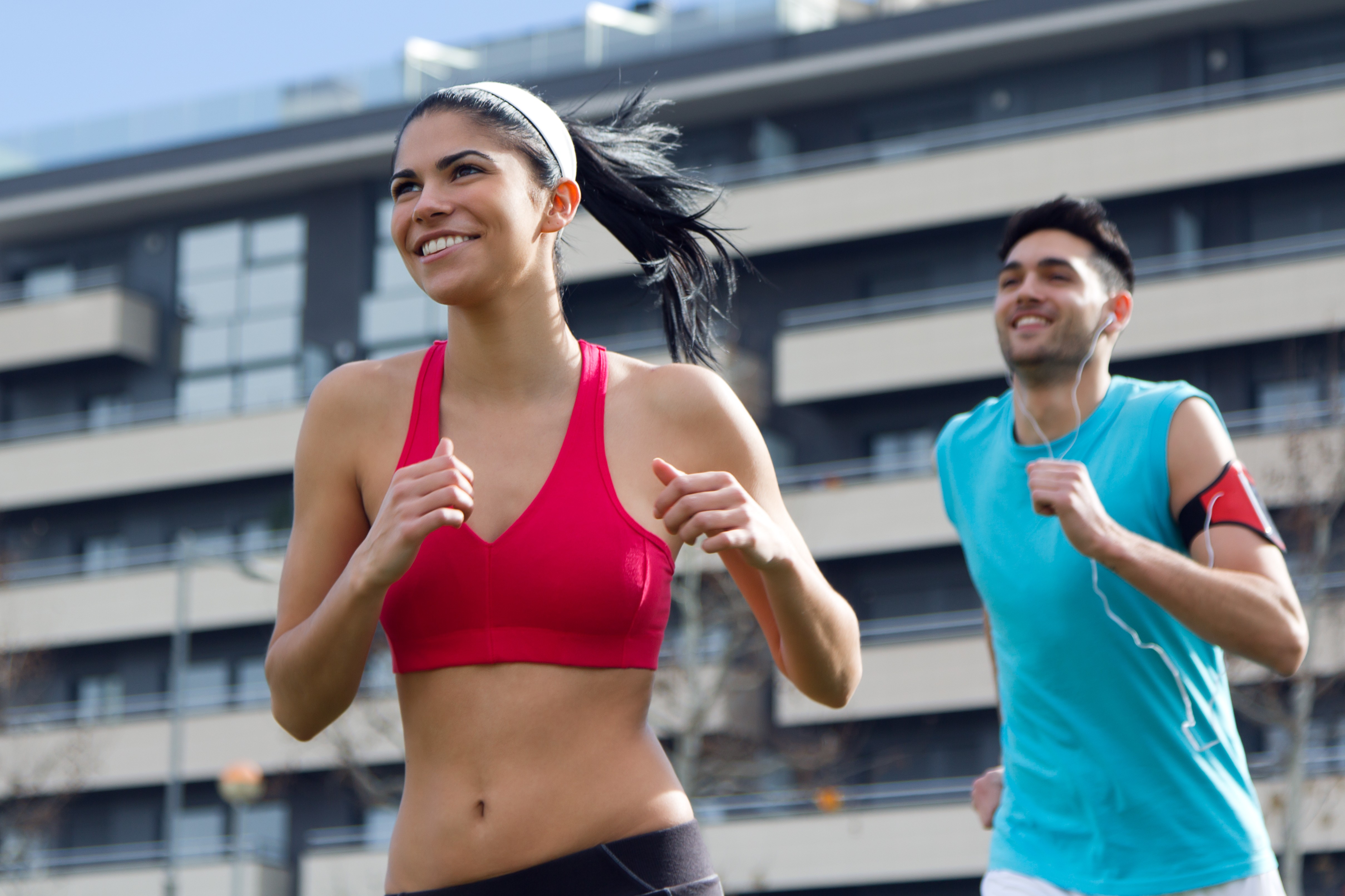 Mulher sorridente de top esportivo vermelho e faixa branca na cabeça correndo ao lado de um homem de camiseta azul com fone de ouvido, ambos praticando exercícios ao ar livre em frente a um prédio moderno