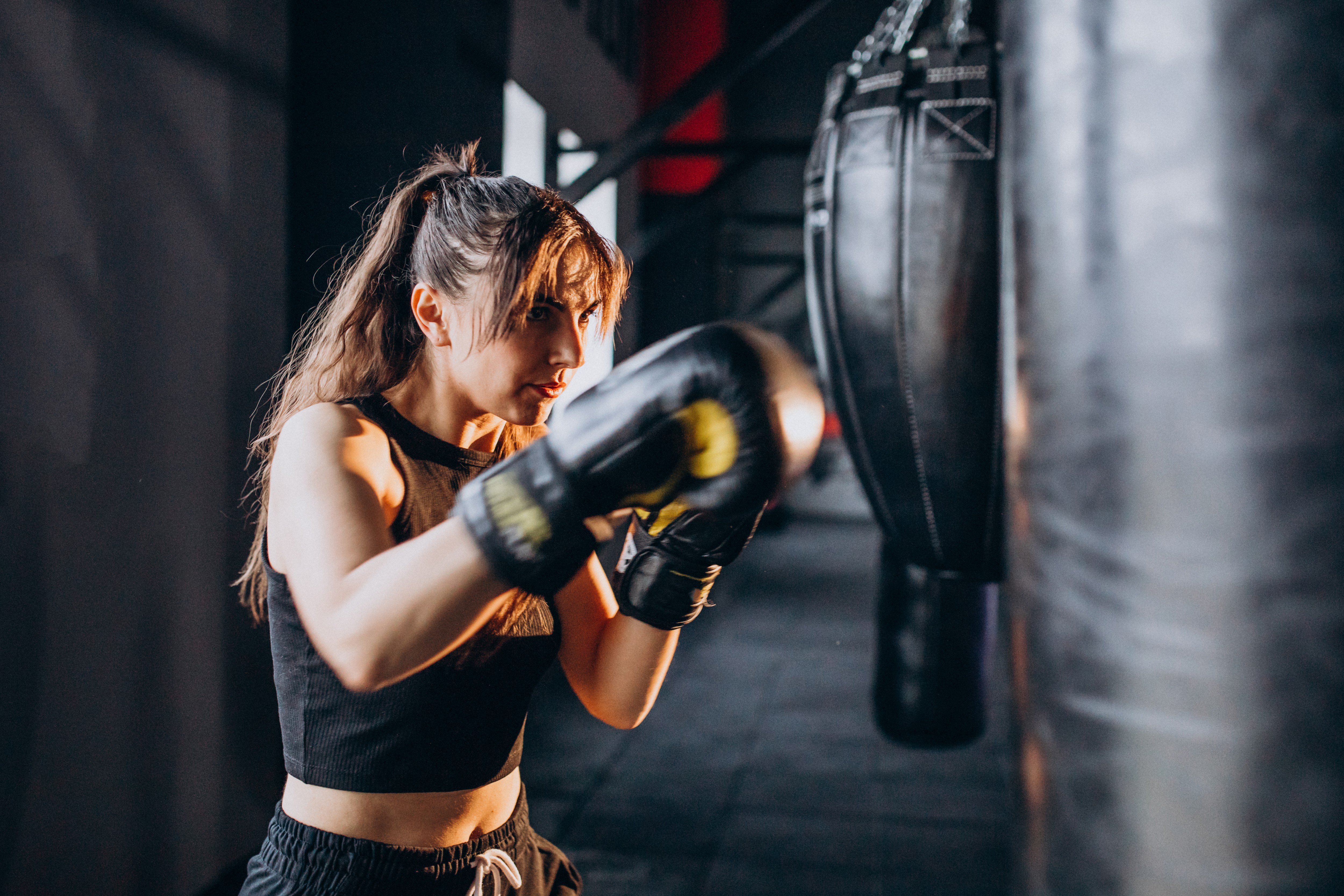 Mulher jovem, com cabelo castanho preso em rabo de cavalo, usando luvas de boxe pretas e roupa esportiva escura, treinando com um saco de pancadas em uma academia