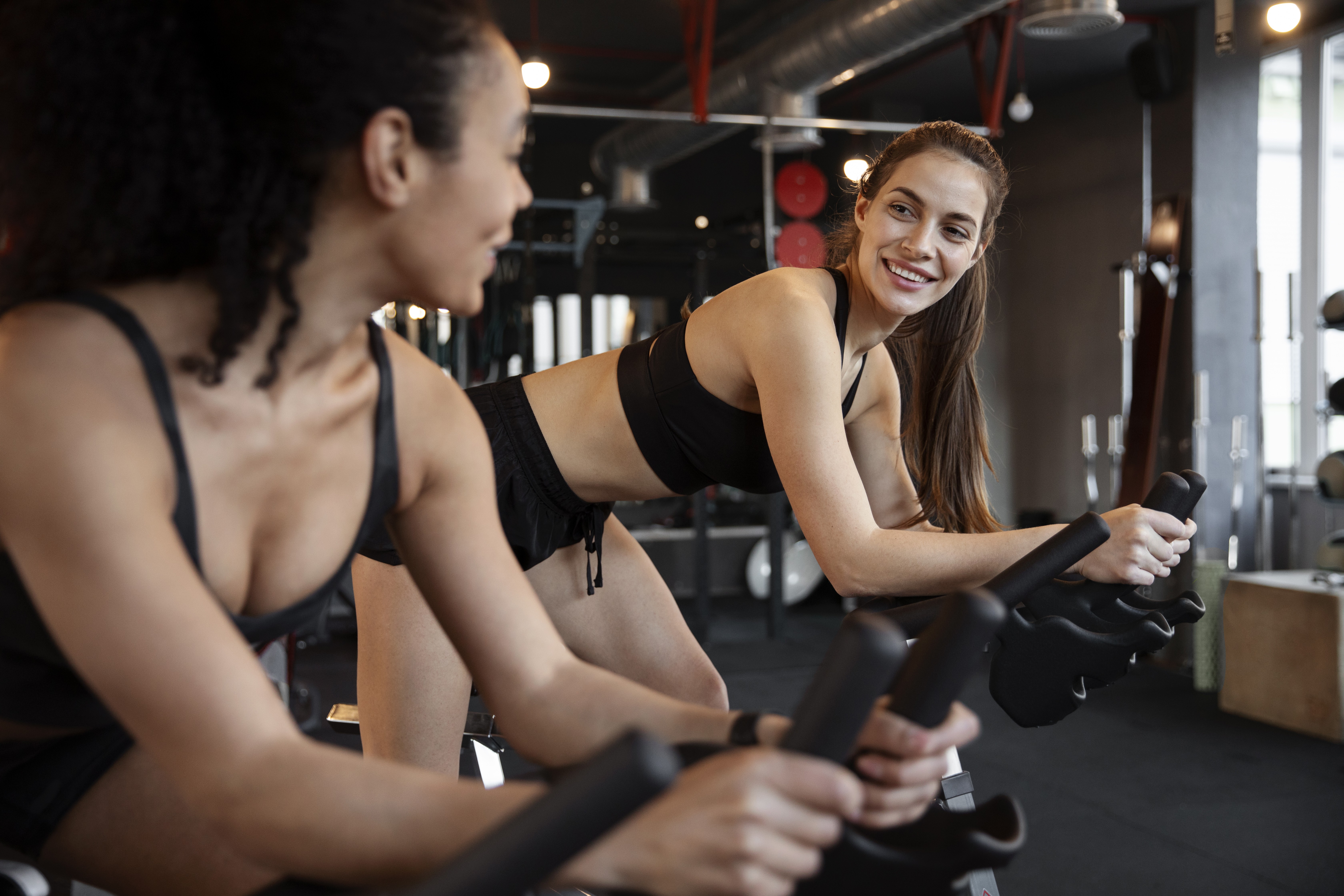 Duas mulheres jovens, vestindo roupas esportivas pretas, sorriem enquanto se exercitam em bicicletas ergométricas na academia