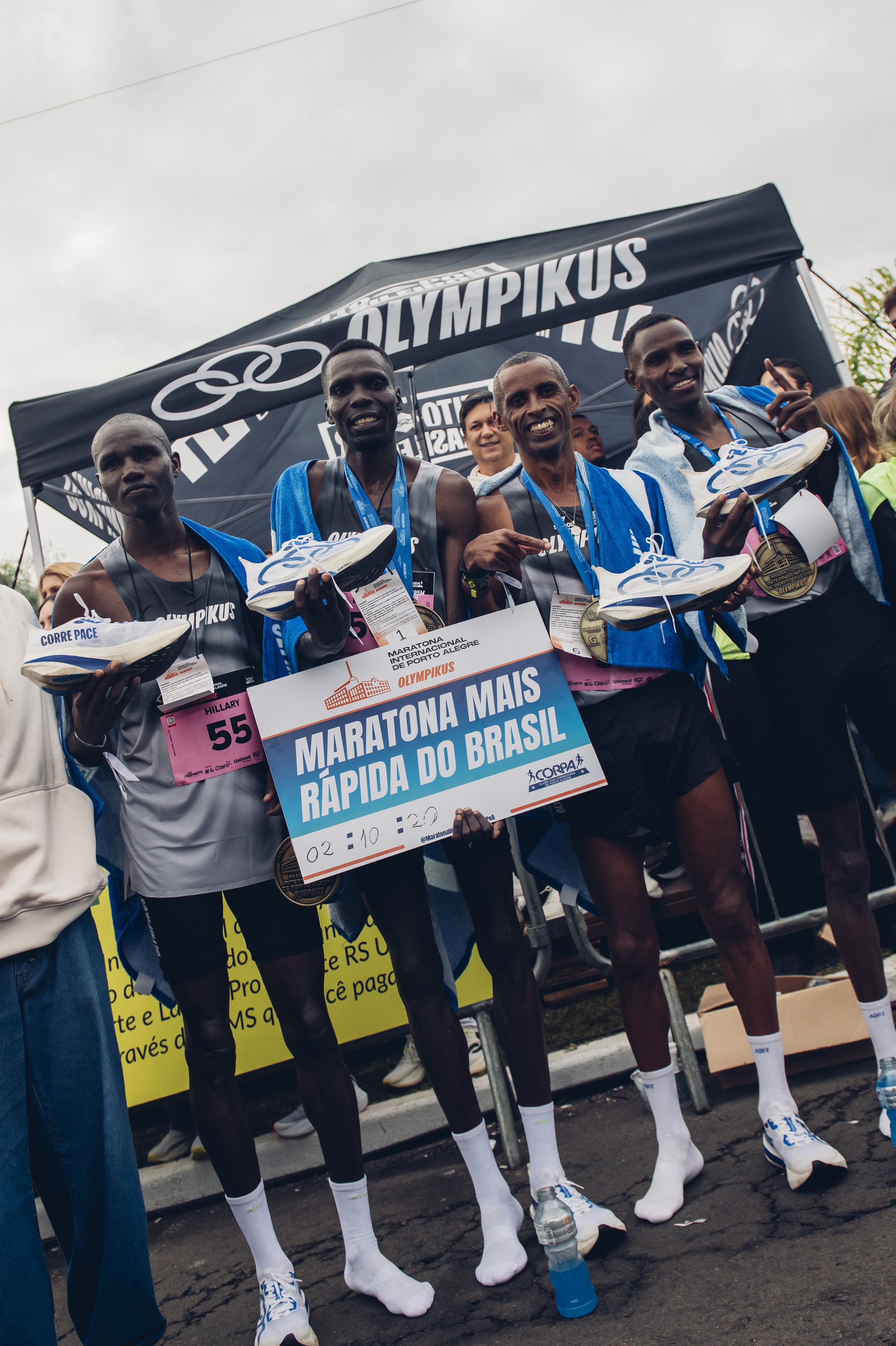 Quatro corredores negros, sorrindo e segurando t&ecirc;nis de corrida brancos e azuis, medalhas e um cartaz azul com 