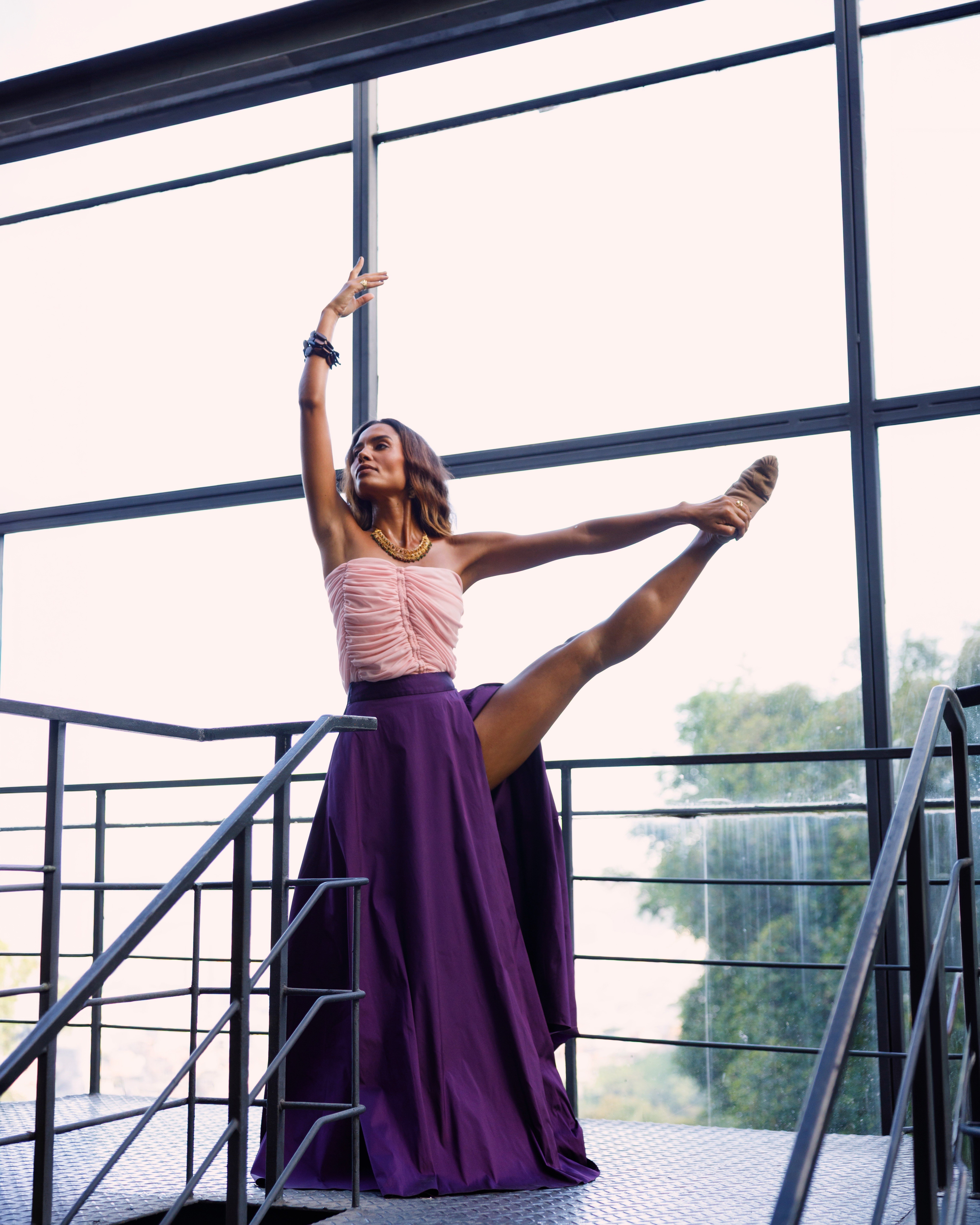 Mulher de pele morena, cabelo castanho ondulado, top rosa drapeado e saia longa roxa, em pose de dan&ccedil;a com uma perna esticada para o lado e um bra&ccedil;o levantado, em uma escada met&aacute;lica com janelas grandes ao fundo
