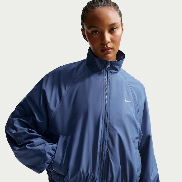 Mulher de pele escura com cabelo tran&ccedil;ado, vestindo uma jaqueta azul-marinho folgada com z&iacute;per e gola alta, e o logo branco da Nike no peito esquerdo. Ela tem a m&atilde;o esquerda no bolso e olha diretamente para a frente. O fundo &eacute; branco