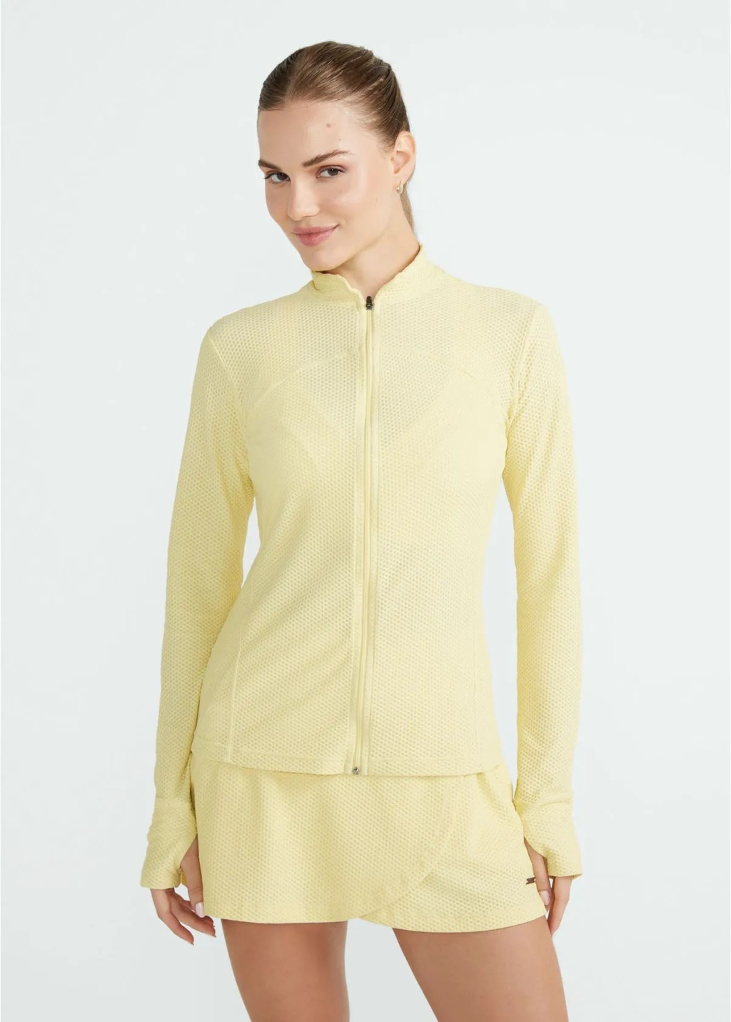 Mulher jovem, de pele clara e cabelo castanho preso, sorri levemente enquanto veste um conjunto esportivo amarelo-claro texturizado, composto por jaqueta de z&iacute;per e saia-short, em fundo branco