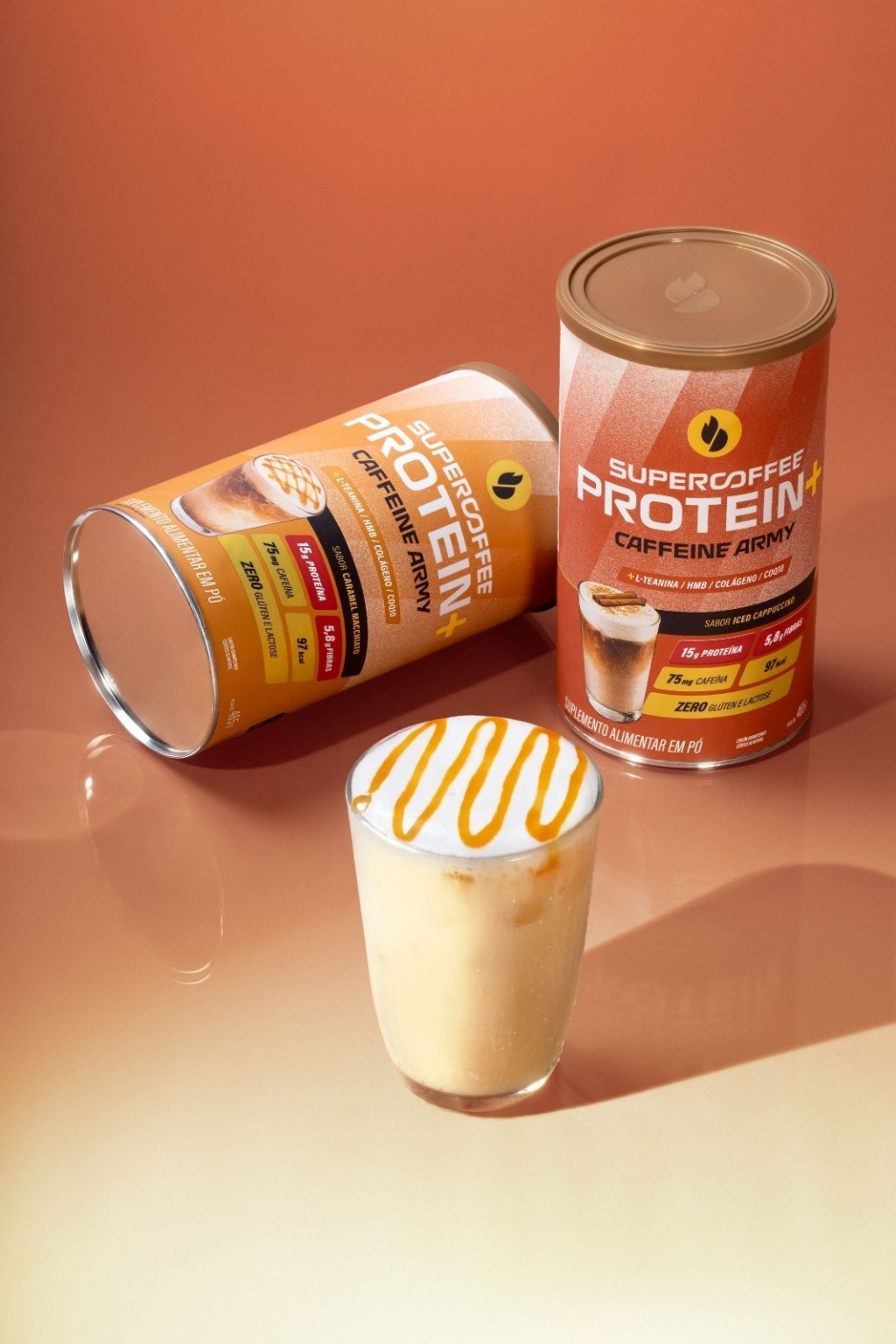 Duas latas de Supercoffee Protein Caffeine Army, uma em p&eacute; e outra deitada, com um copo de bebida cremosa com calda de caramelo ao lado, sobre um fundo laranja