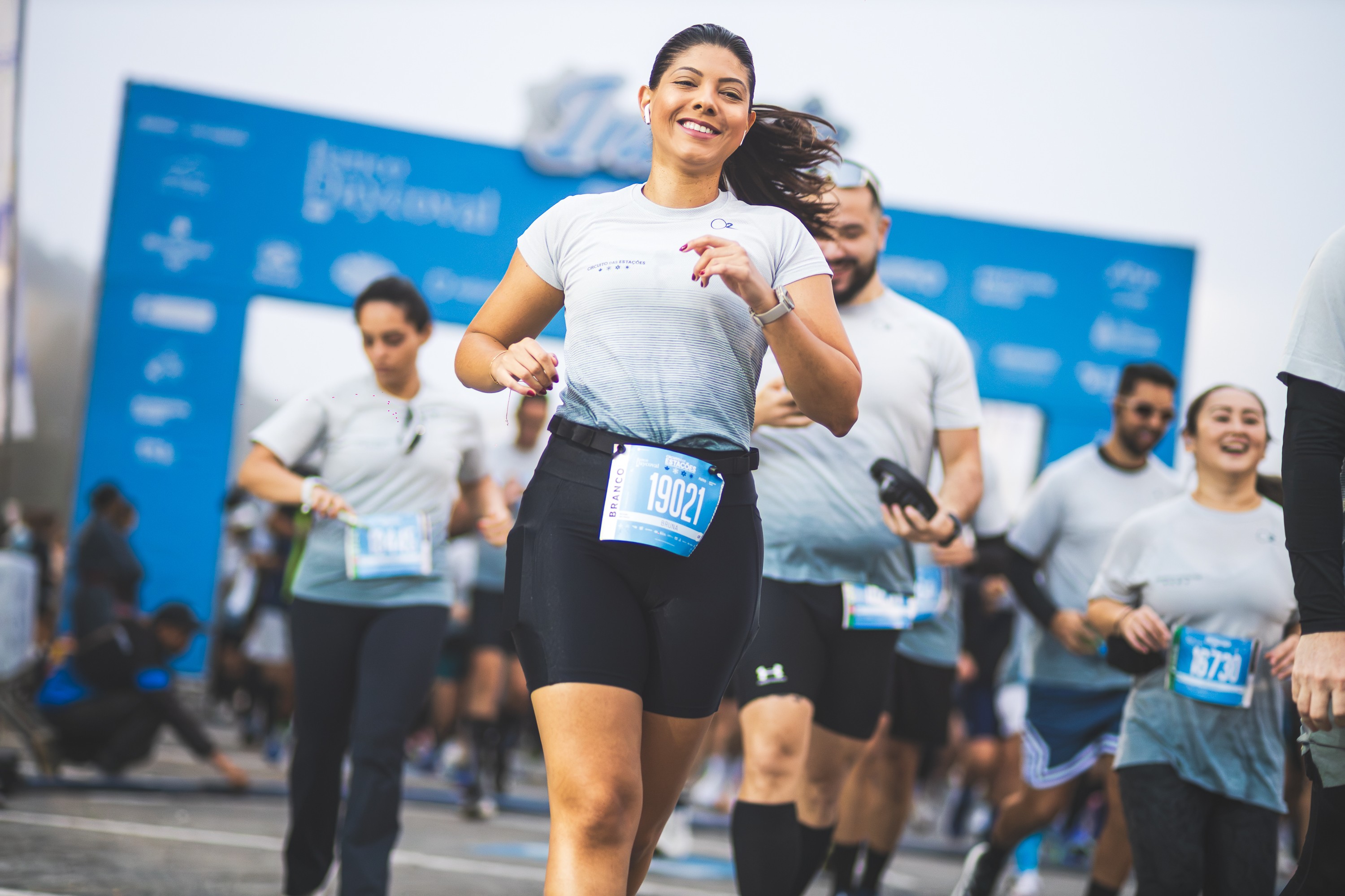 Mulher sorridente de camiseta cinza e shorts pretos correndo em uma maratona, com o n&uacute;mero de peito 19021. Outros corredores e um arco azul de largada/chegada desfocado ao fundo.