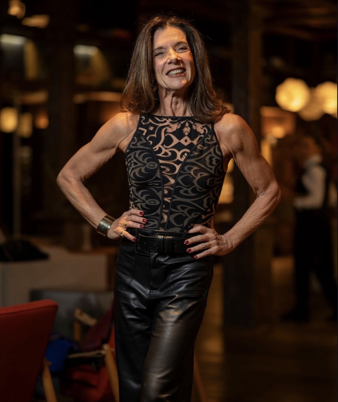 Mulher de meia-idade, cabelos castanhos e sorriso largo, vestindo blusa preta com estampa geom&eacute;trica e cal&ccedil;a de couro preta, posa com as m&atilde;os na cintura em ambiente interno com ilumina&ccedil;&atilde;o baixa