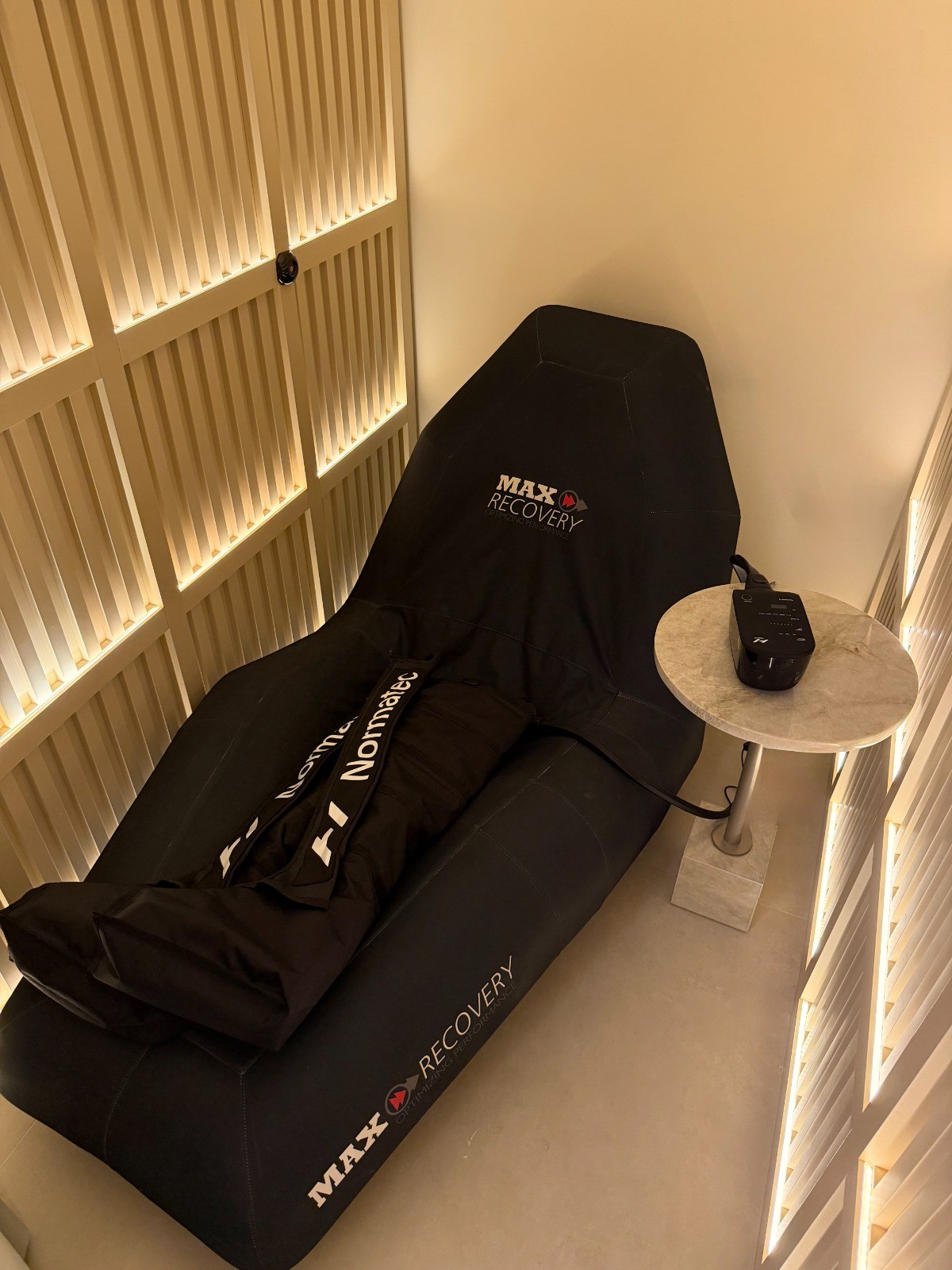 Poltrona reclin&aacute;vel preta da marca Max Recovery com botas de compress&atilde;o Normatec, ao lado de uma mesa de cabeceira redonda de m&aacute;rmore com um aparelho eletr&ocirc;nico preto, em um ambiente com paredes de ripas de madeira iluminadas por luzes indiretas