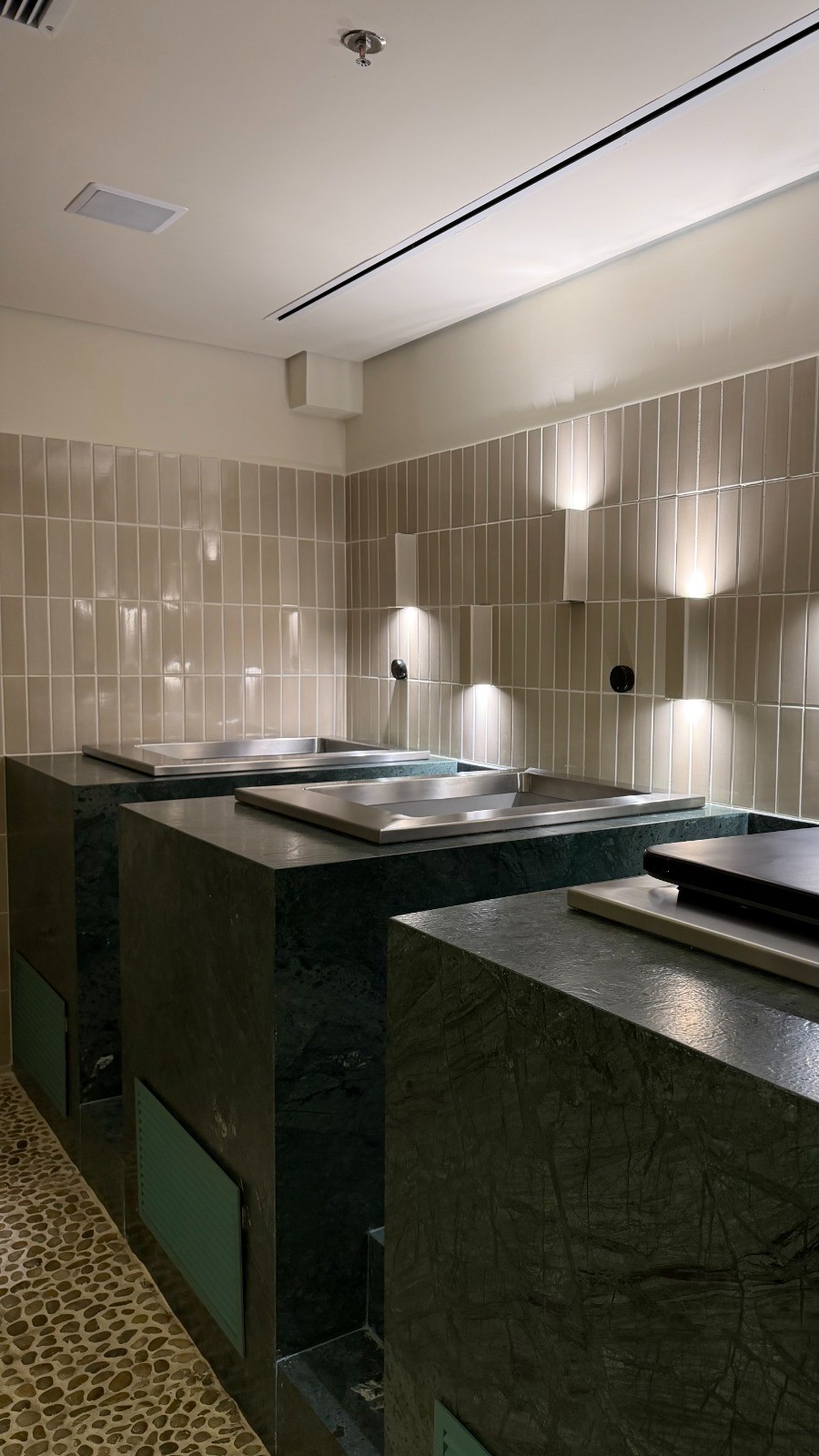 Ambiente interno com tr&ecirc;s bancadas de m&aacute;rmore verde escuro e cubas de inox. A parede &eacute; revestida com azulejos bege claros e h&aacute; lumin&aacute;rias de parede que emitem luz para cima e para baixo. O ch&atilde;o &eacute; de pedras claras e o teto tem ilumina&ccedil;&atilde;o linear e um sprinkler.