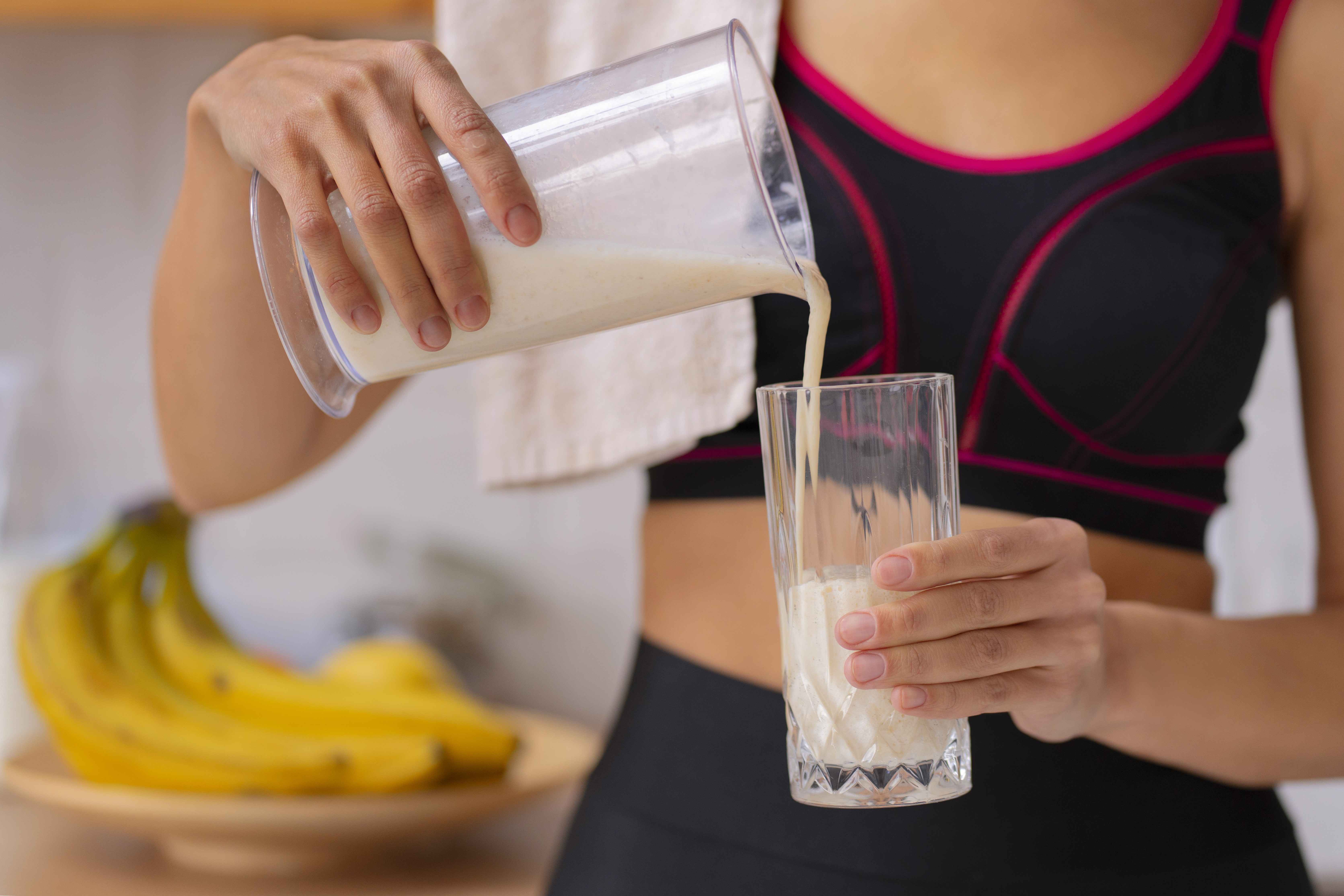 Mulher em top esportivo preto e rosa despeja vitamina de liquidificador em copo de vidro, com bananas em fruteira ao fundo