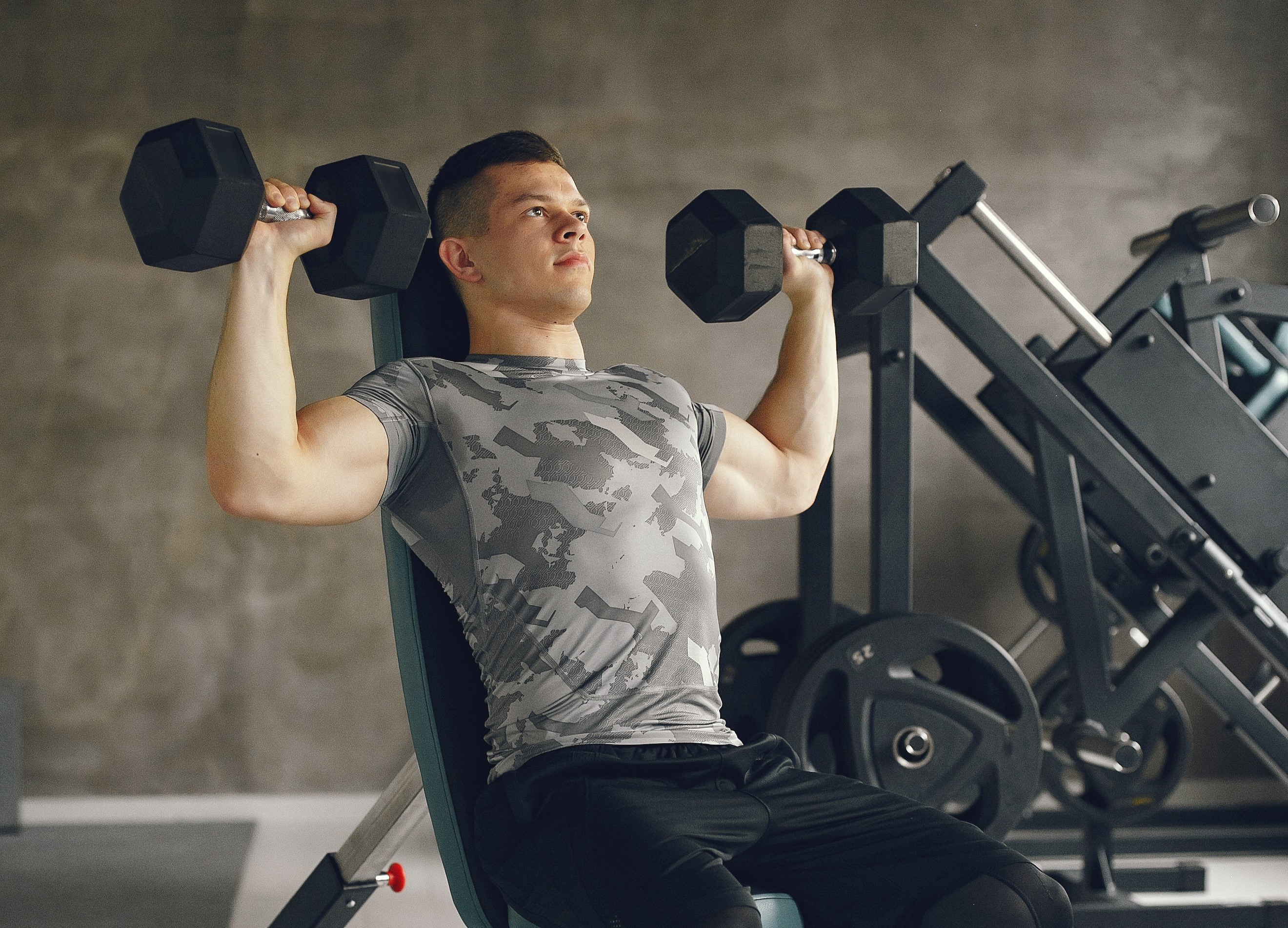 Homem jovem, branco, com cabelo curto e escuro, usando camiseta cinza camuflada e cal&ccedil;a preta, sentado em um banco de academia, levantando halteres pretos com os bra&ccedil;os flexionados acima da cabe&ccedil;a, em um ambiente de academia com equipamentos de muscula&ccedil;&atilde;o ao fundo