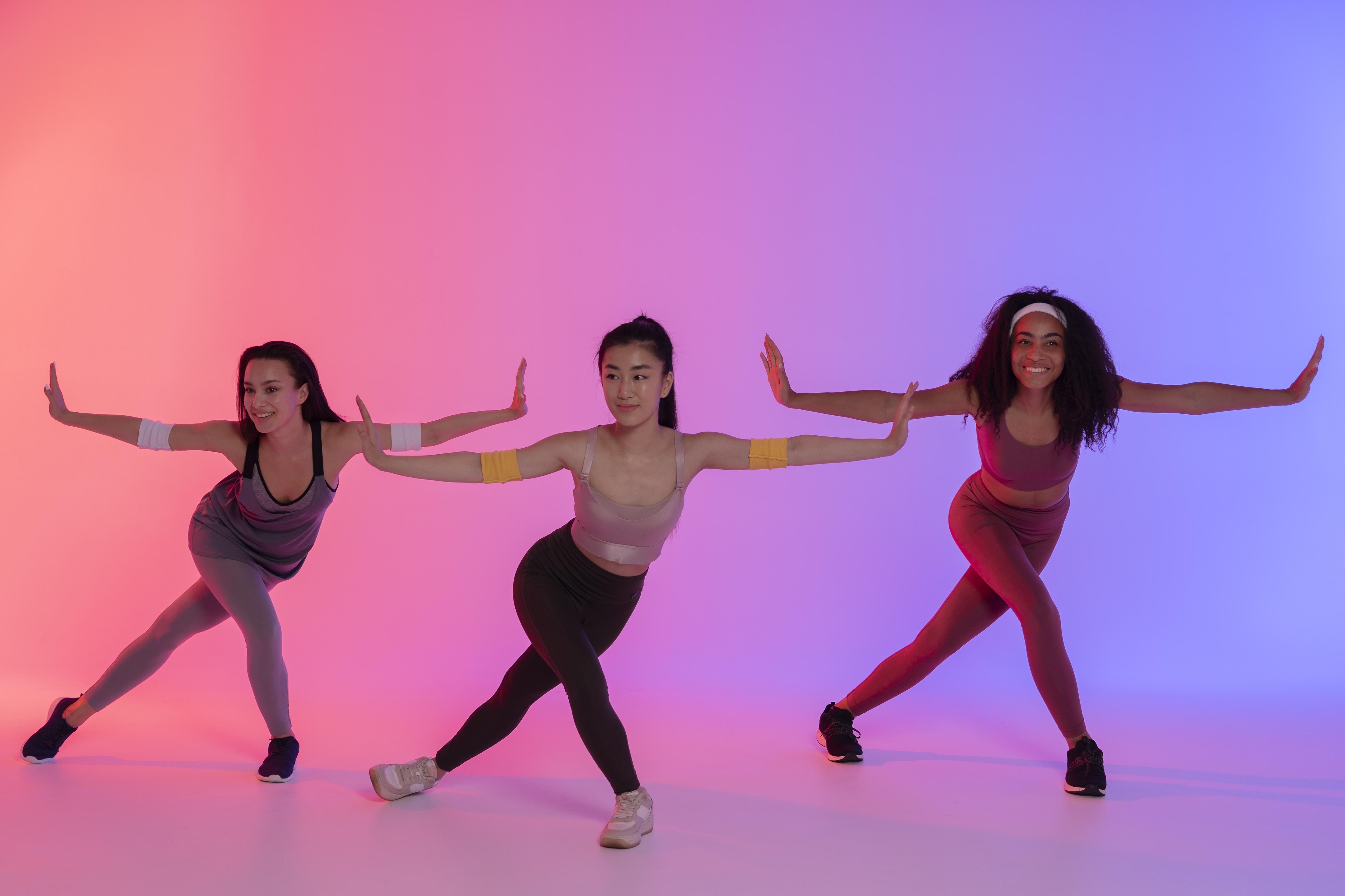 Tr&ecirc;s mulheres jovens, de etnias diversas, sorriem enquanto dan&ccedil;am em um est&uacute;dio com ilumina&ccedil;&atilde;o neon rosa e azul. Elas usam roupas esportivas e est&atilde;o com os bra&ccedil;os estendidos, palmas das m&atilde;os para frente, e pernas flexionadas em uma pose din&acirc;mica