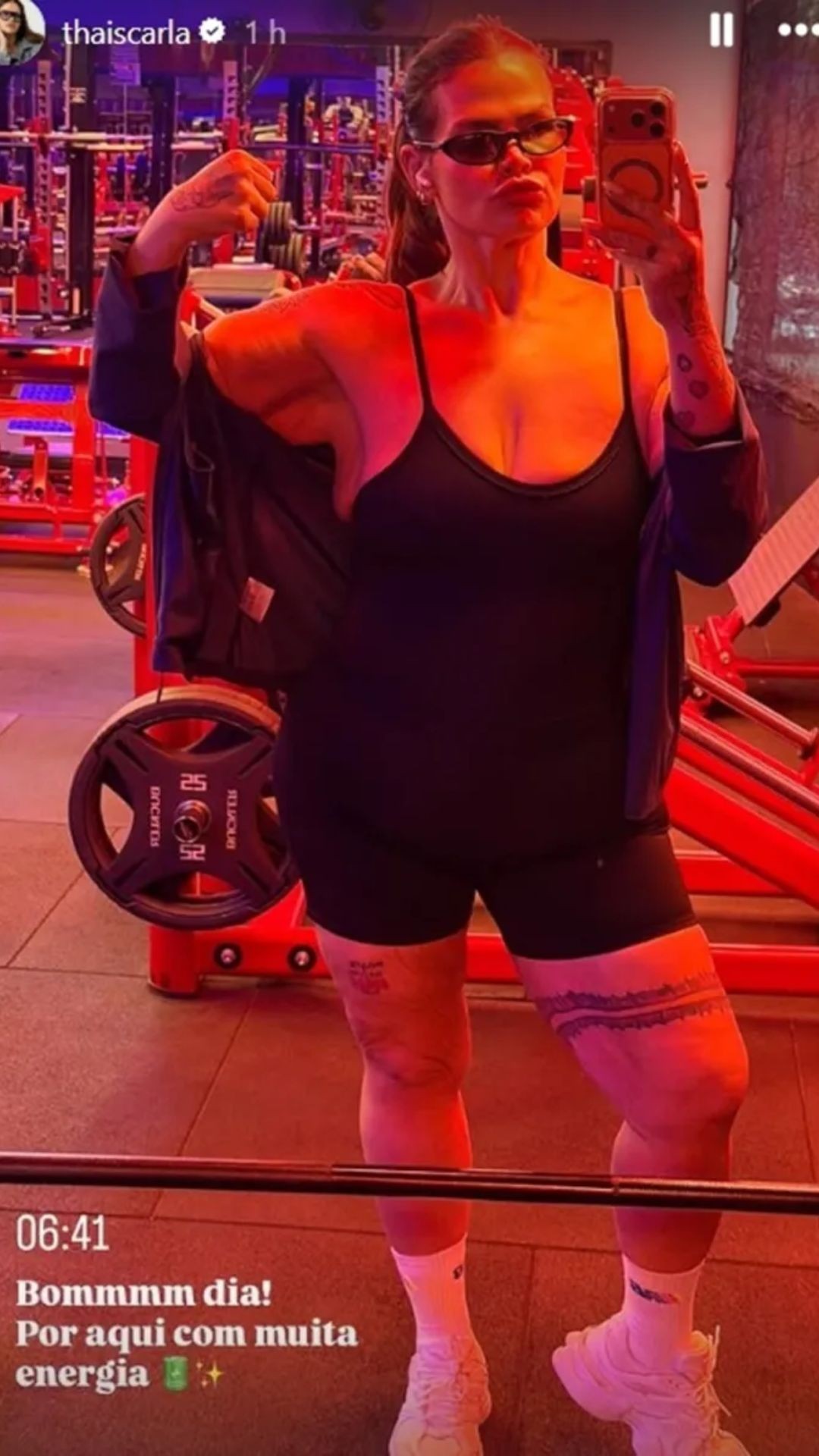 Thais Carla posa em selfie de corpo inteiro na academia, vestindo top e shorts pretos, jaqueta azul escura e &oacute;culos de sol. Ela faz bico e flexiona o bra&ccedil;o direito, exibindo tatuagens nos bra&ccedil;os e pernas. O ambiente tem ilumina&ccedil;&atilde;o avermelhada e equipamentos de muscula&ccedil;&atilde;o vermelhos ao fundo. No canto inferior esquerdo, texto branco diz 06:41 Bommmm dia! Por aqui com muita energia.