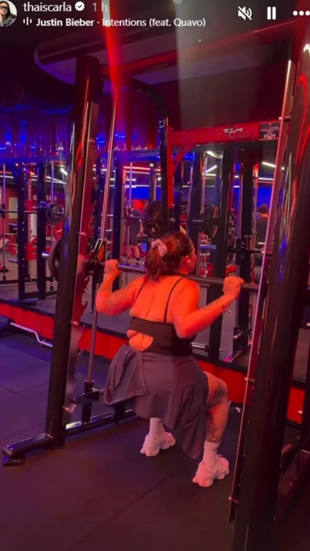 Mulher de costas, com top preto e cal&ccedil;a cinza, agacha segurando uma barra de peso em uma m&aacute;quina de academia com ilumina&ccedil;&atilde;o vermelha e azul