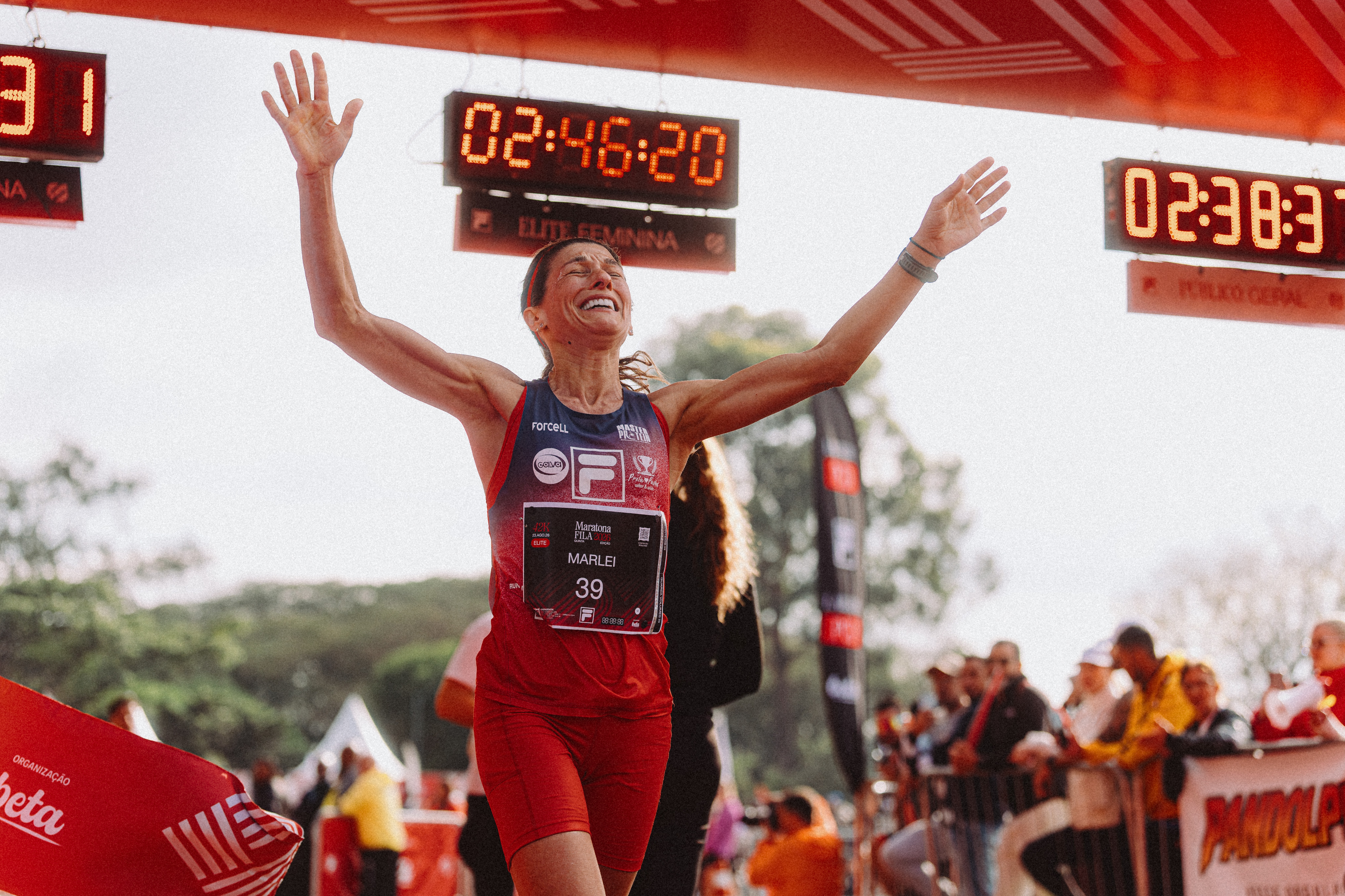 Mulher atleta com camiseta azul e vermelha, n&uacute;mero 39, cruza a linha de chegada de uma corrida com os bra&ccedil;os erguidos em celebra&ccedil;&atilde;o. Ela sorri, com o rosto virado para cima, sob um placar digital que marca 02:46:20. Pessoas assistem ao fundo