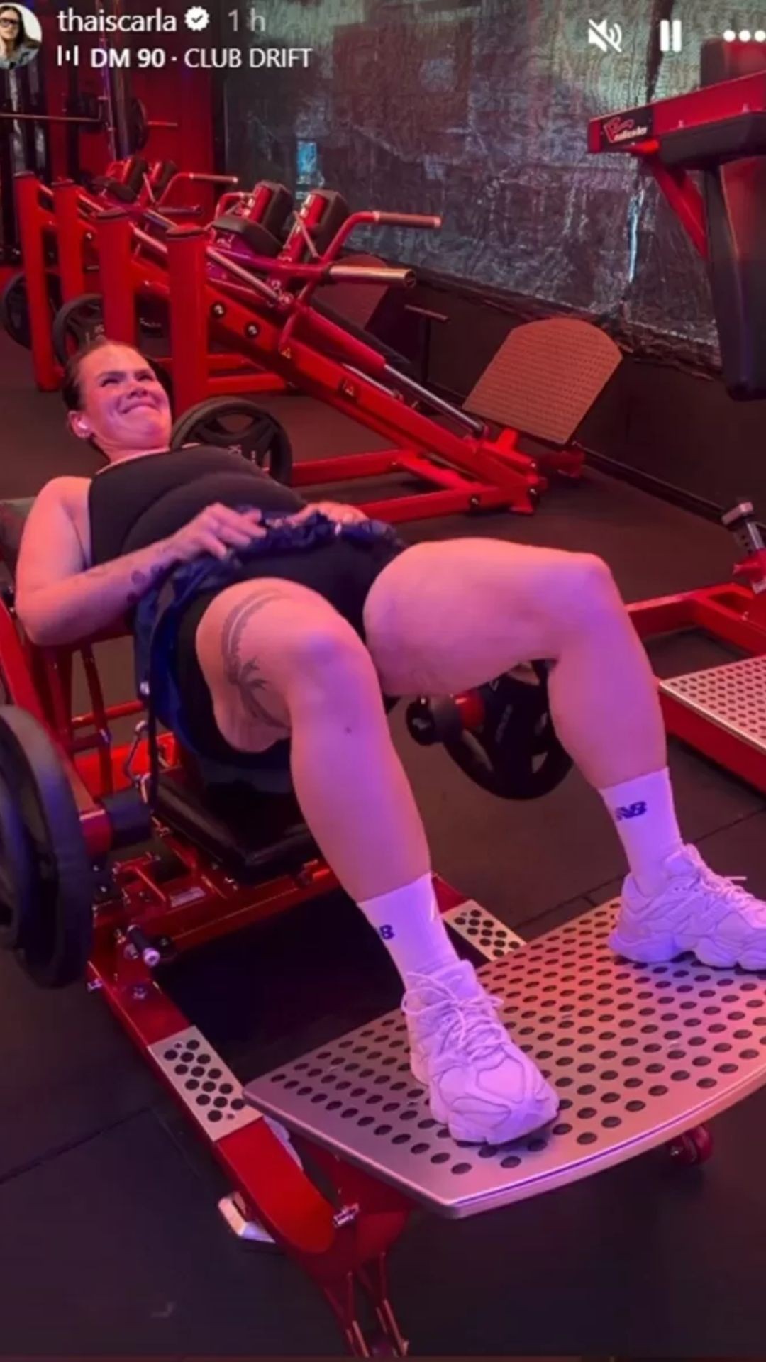 Mulher deitada em aparelho de academia vermelho, fazendo exerc&iacute;cio para as pernas, com express&atilde;o de esfor&ccedil;o e sorriso. Veste top preto, shorts escuro e t&ecirc;nis brancos, exibindo tatuagens na coxa.