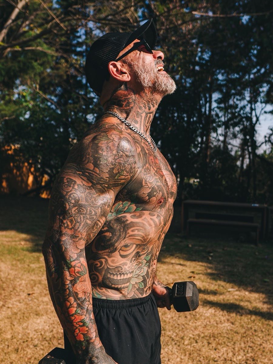 Homem musculoso e tatuado, com barba grisalha, bon&eacute; preto e &oacute;culos escuros, levanta o rosto para o c&eacute;u enquanto segura um halter em cada m&atilde;o, exercitando-se ao ar livre em um dia ensolarado