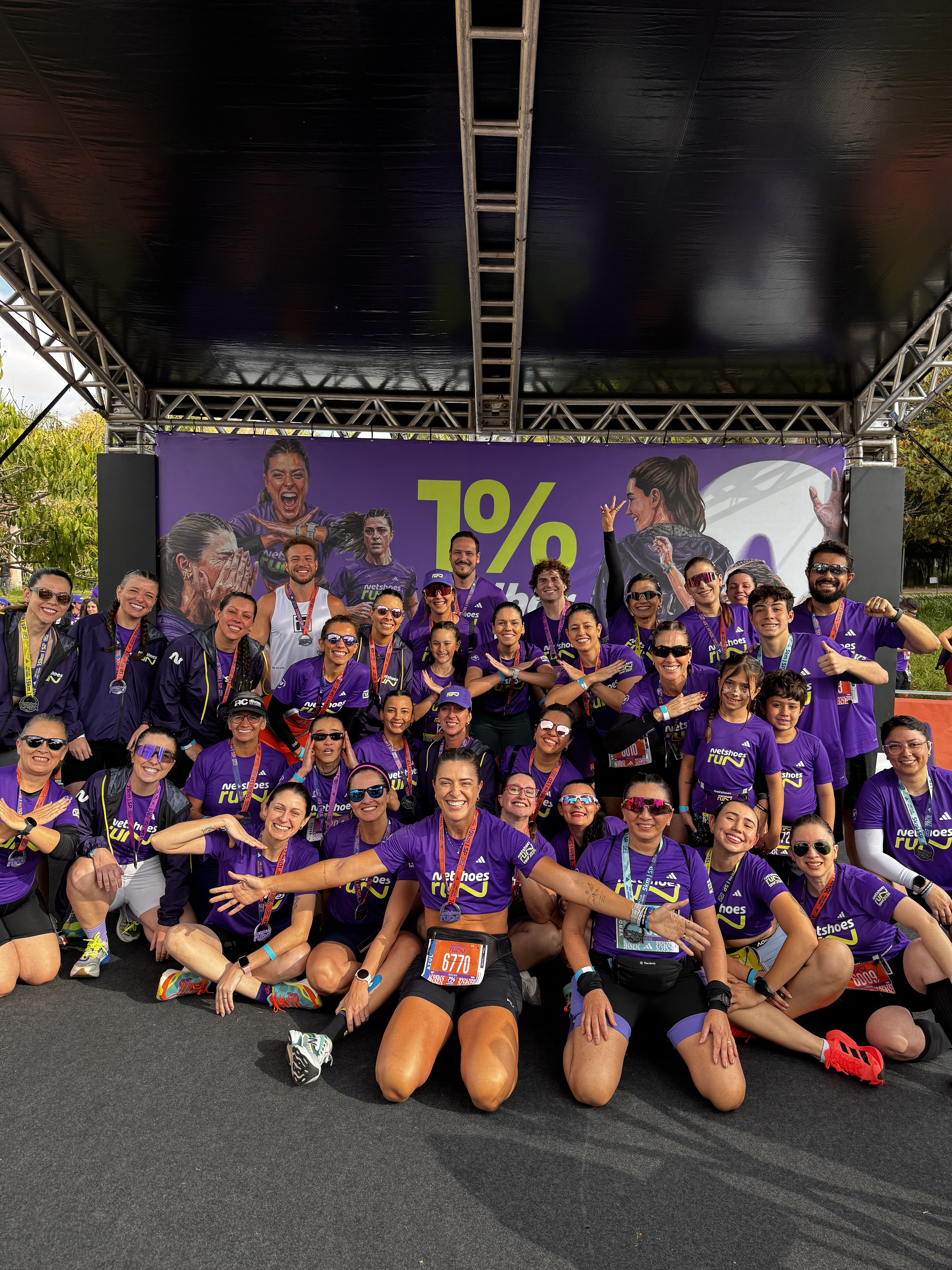 Um grupo de cerca de 30 corredores, a maioria mulheres, sorriem para a c&acirc;mera em frente a um banner roxo com a frase 1%. Todos vestem camisetas roxas com o logo Netshoes Run e muitos exibem medalhas no pesco&ccedil;o. Alguns est&atilde;o agachados, outros em p&eacute;, com express&otilde;es de alegria e satisfa&ccedil;&atilde;o ap&oacute;s a corrida