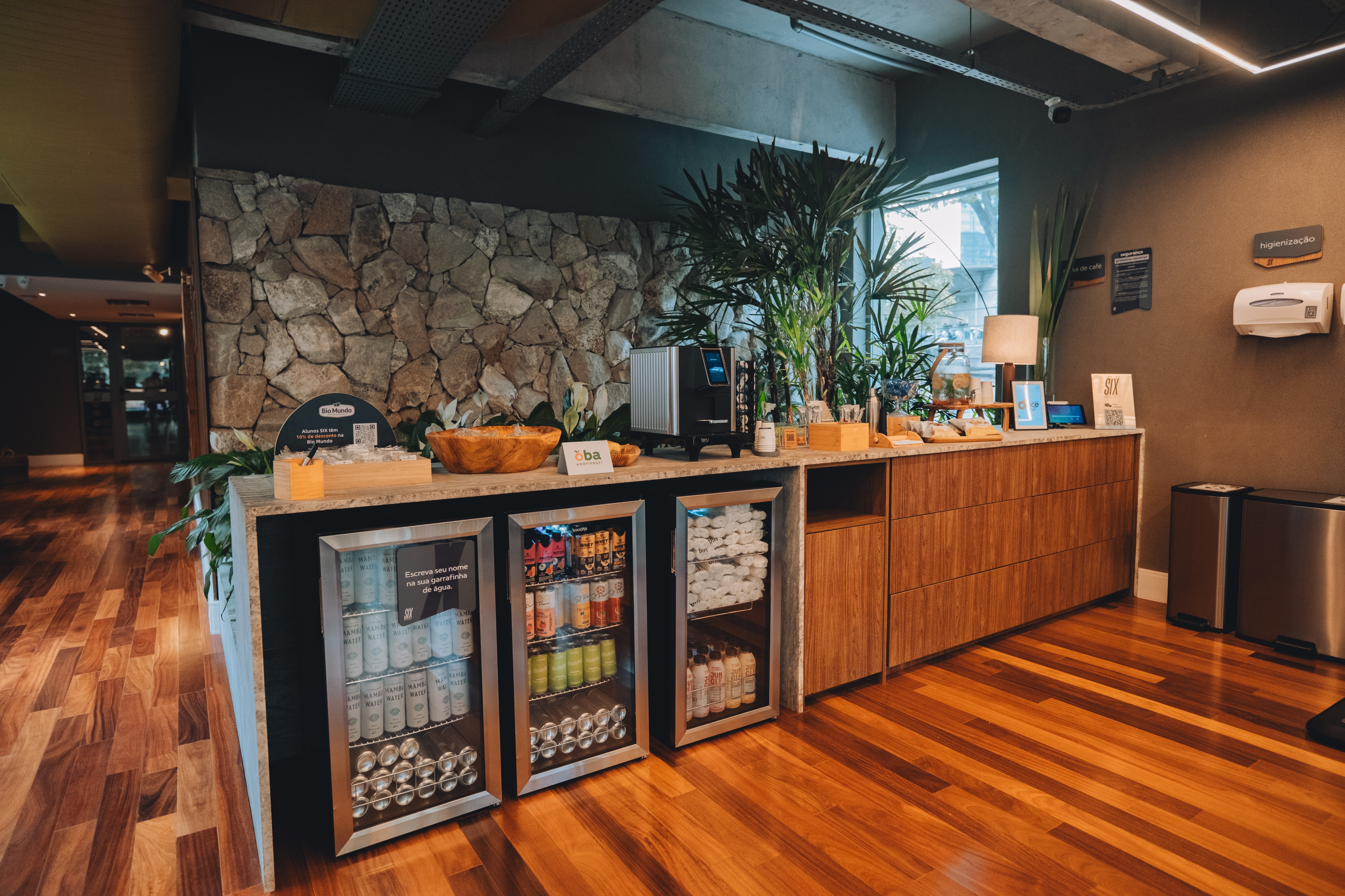 Balc&atilde;o de madeira com tr&ecirc;s frigobares de vidro, m&aacute;quina de caf&eacute;, tigela de madeira com frutas, plantas e lixeiras de inox em ambiente com parede de pedras e piso de madeira