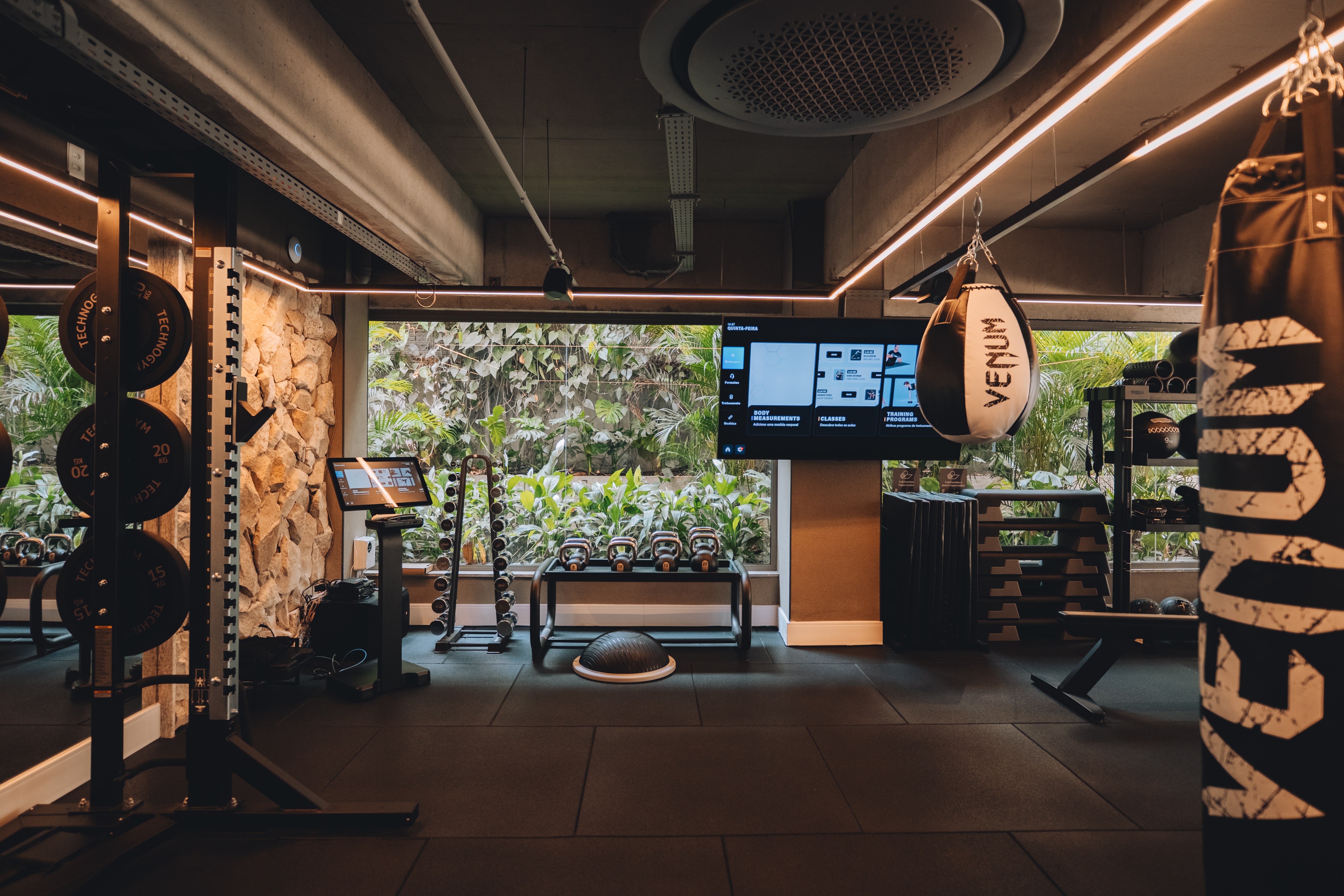 Academia moderna com piso emborrachado, equipamentos de muscula&ccedil;&atilde;o e pesos. &Agrave; esquerda, anilhas em um rack; ao centro, halteres e kettlebells. Ao fundo, uma grande janela revela vegeta&ccedil;&atilde;o exuberante. Uma TV exibe informa&ccedil;&otilde;es de treino e um saco de pancadas preto e branco com a marca Venum est&aacute; pendurado &agrave; direita