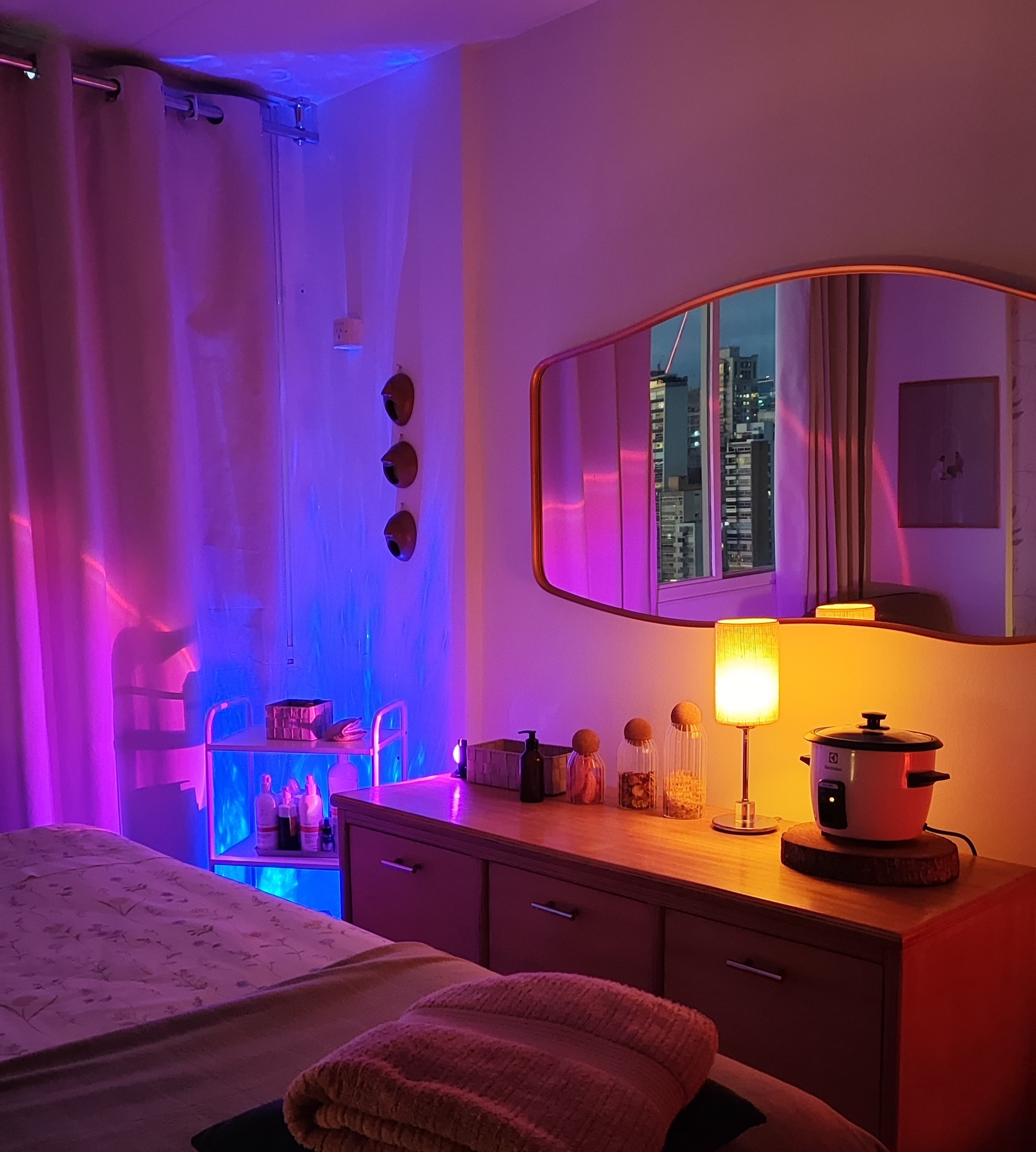 Ambiente de spa ou massagem com ilumina&ccedil;&atilde;o ambiente em tons de roxo e azul, uma maca com toalhas dobradas, um espelho refletindo pr&eacute;dios urbanos, e uma mesa de madeira com lumin&aacute;ria, potes de vidro e um aquecedor de toalhas