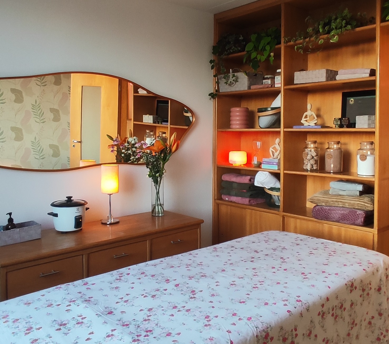 Ambiente de spa com maca coberta por len&ccedil;ol floral, espelho org&acirc;nico, c&ocirc;moda com aquecedor de pedras e lumin&aacute;ria, e estante de madeira com plantas, toalhas e objetos decorativos, como um difusor de aromas aceso